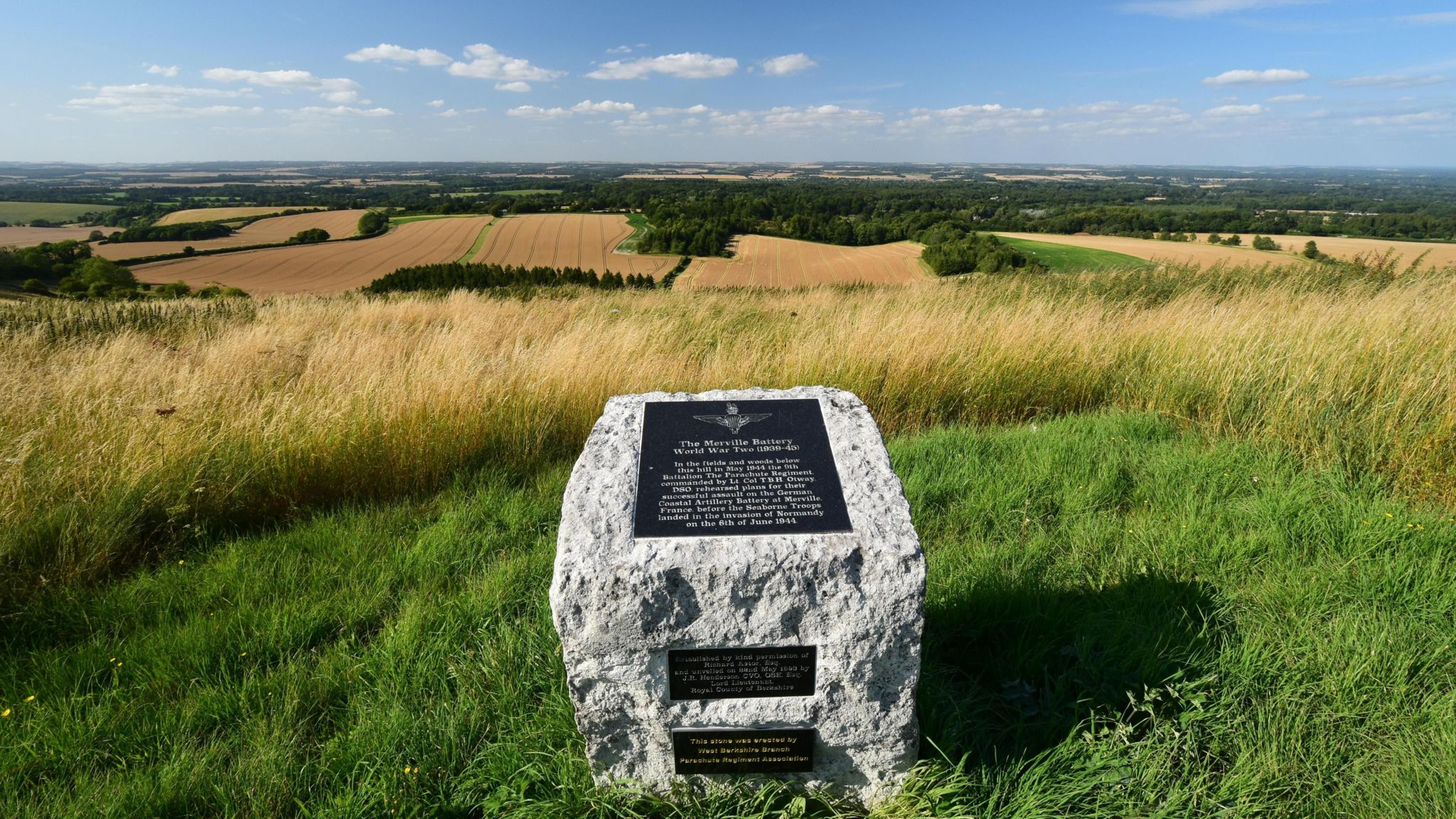 A stone memorial with a brass plaque to the location of a World War II practice battery site stands on a hill. The sky is blue with fluffy clouds. A landscape of wheat fields and trees lies out before it.