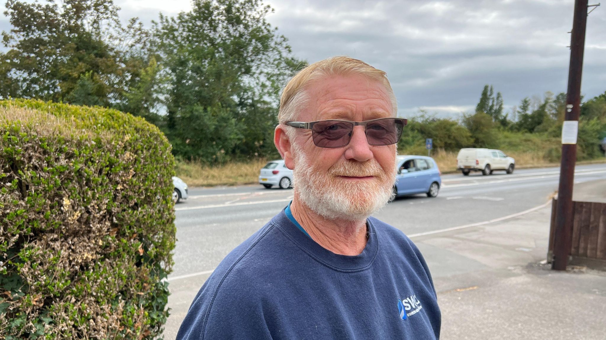 Man looking at camera with A358 road in the background. He has a beard, dark glasses and is wearing a light blue sweatshirt