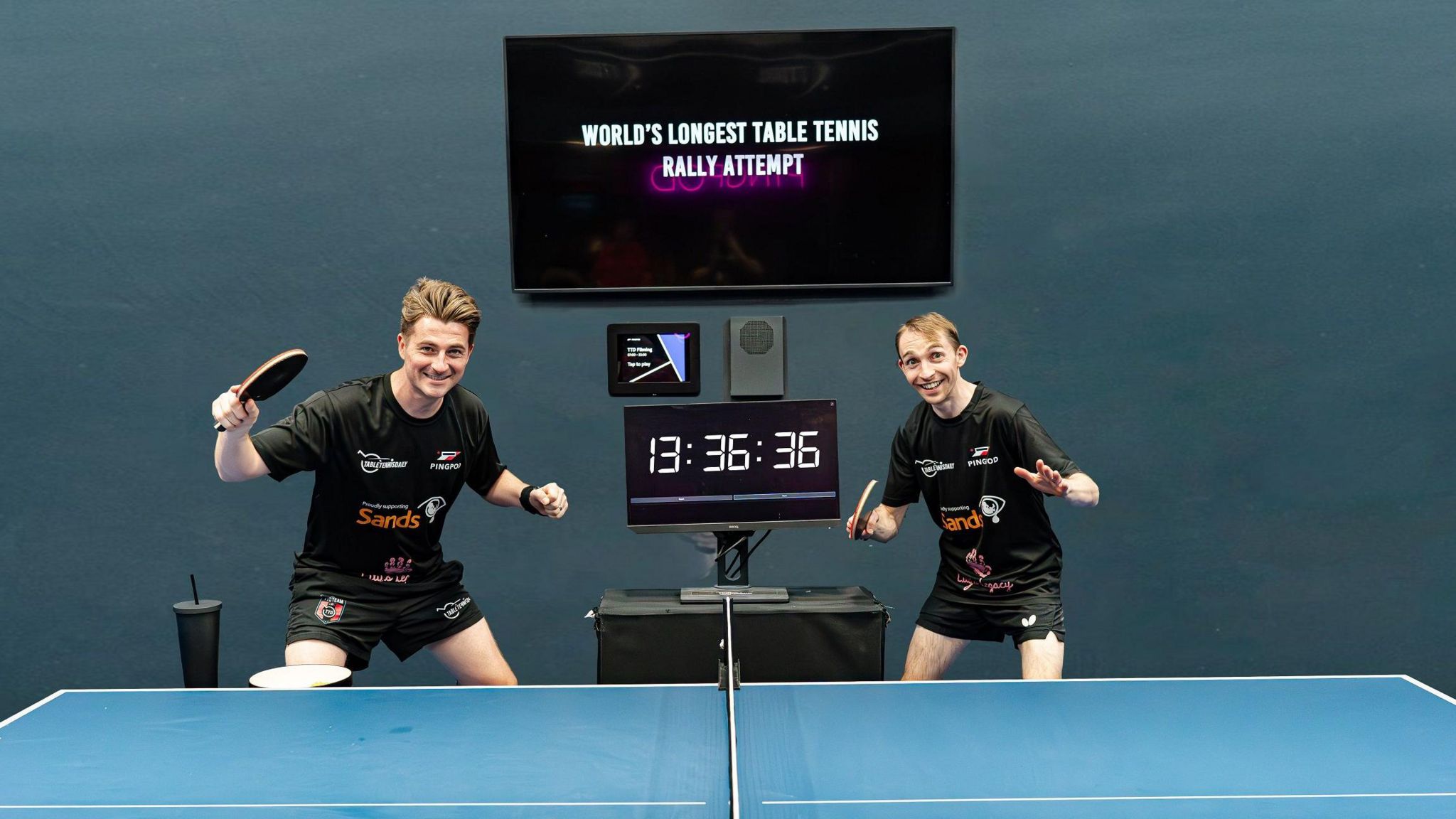 Image of Dan Ives and Lloyd Gregory. They are by a table tennis table and a clock reading 13 hours 36 mins 36 secs. 