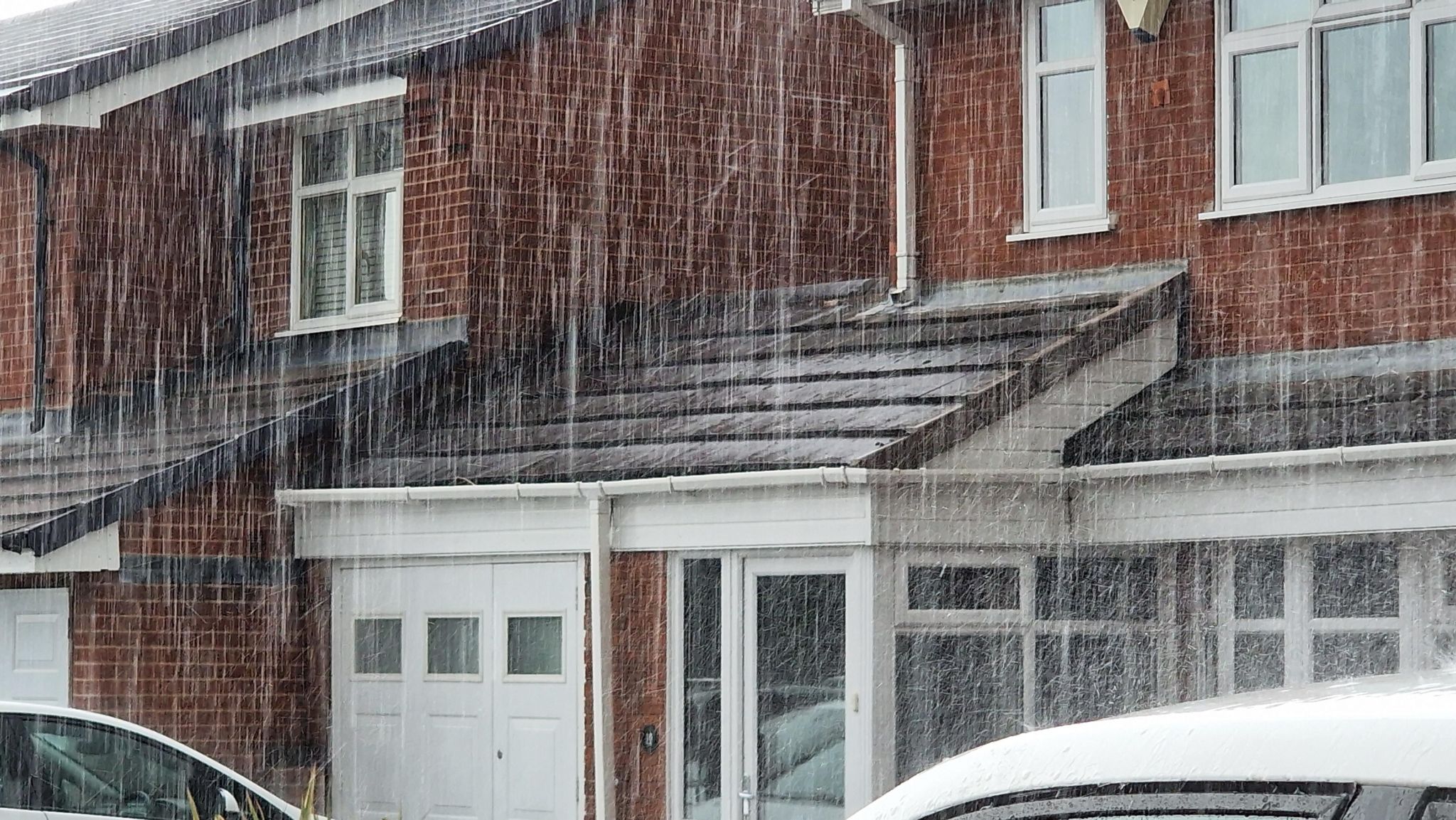 Streaks of very heavy rain falling onto two cars and bouncing off a house roof. The brick house has white doors and window frames. 