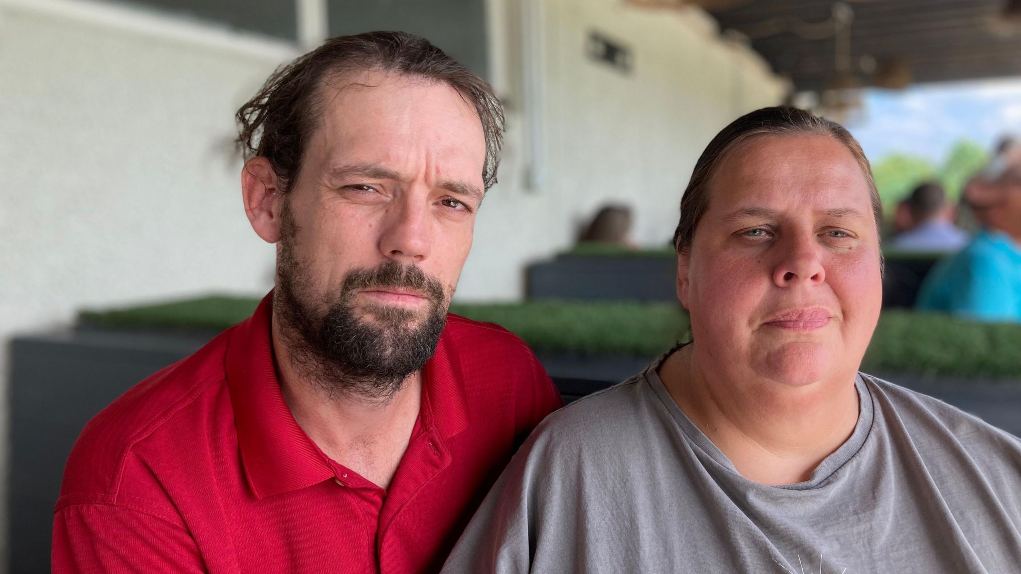 Christopher, left wears a bright red polo shirt and has dark hair and a beard. Jackie wears a grey tshir and has her hair pulled back. Both are unsmiling, with Jackie looking away from the camera .