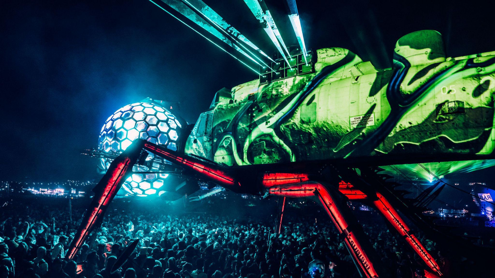 A biomechanical dragonfly lit up in blue and green with thousands of people dancing underneath.