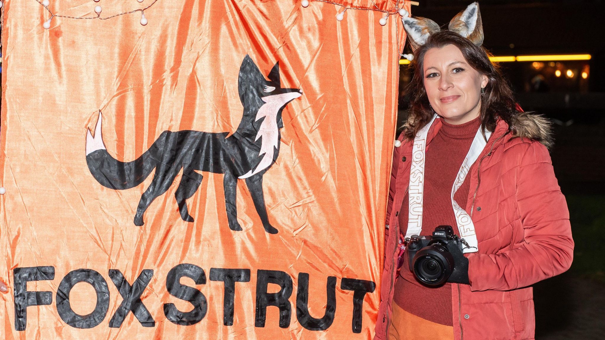 Jayne Jackson, co-founder of Fox Strut, wearing fox ears and a deep red and orange outfit, and holding an orange Fox Strut banner and a camera