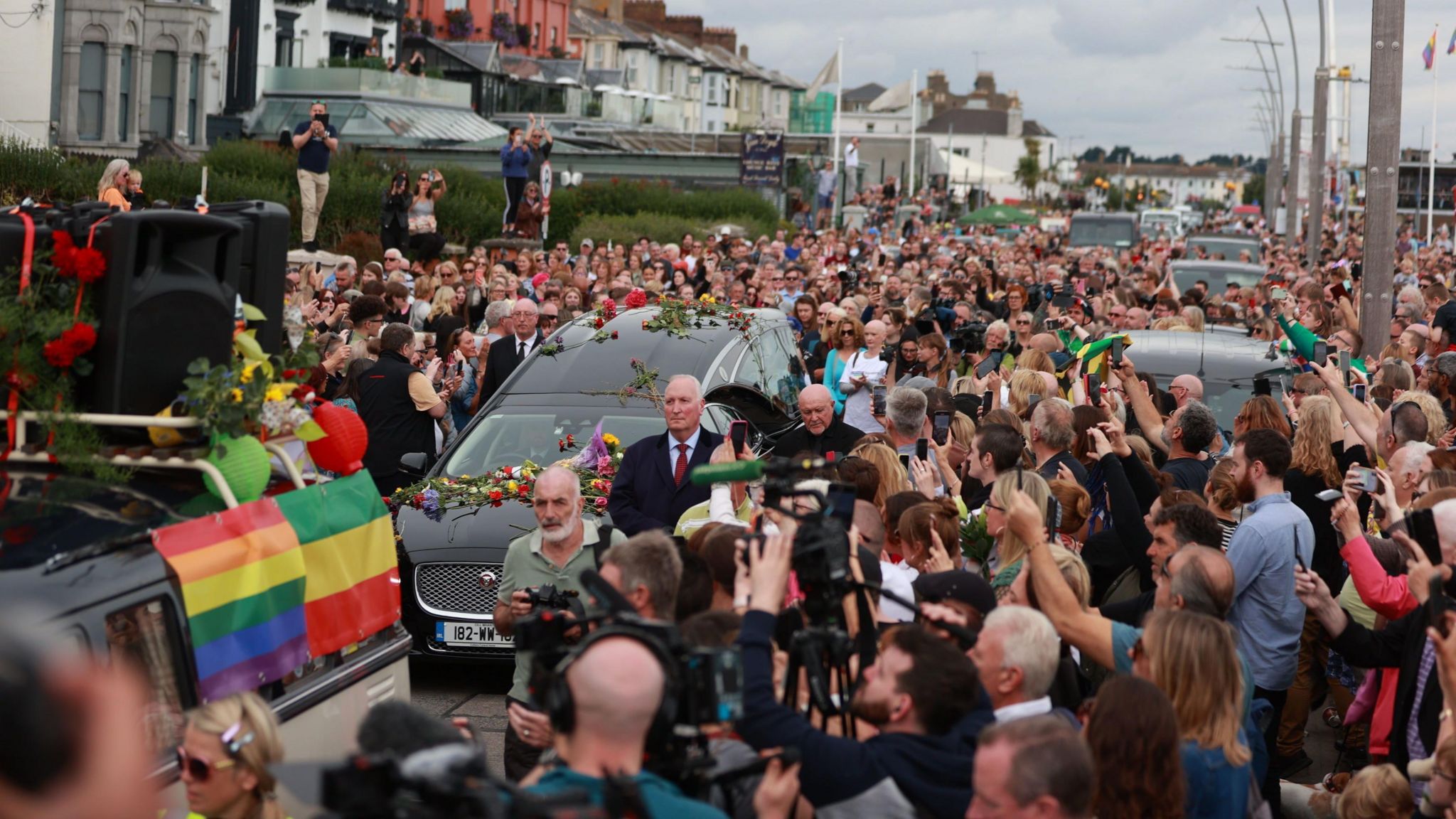 A huddle of people stand on the street as a hearse covered in flowers drives through the street