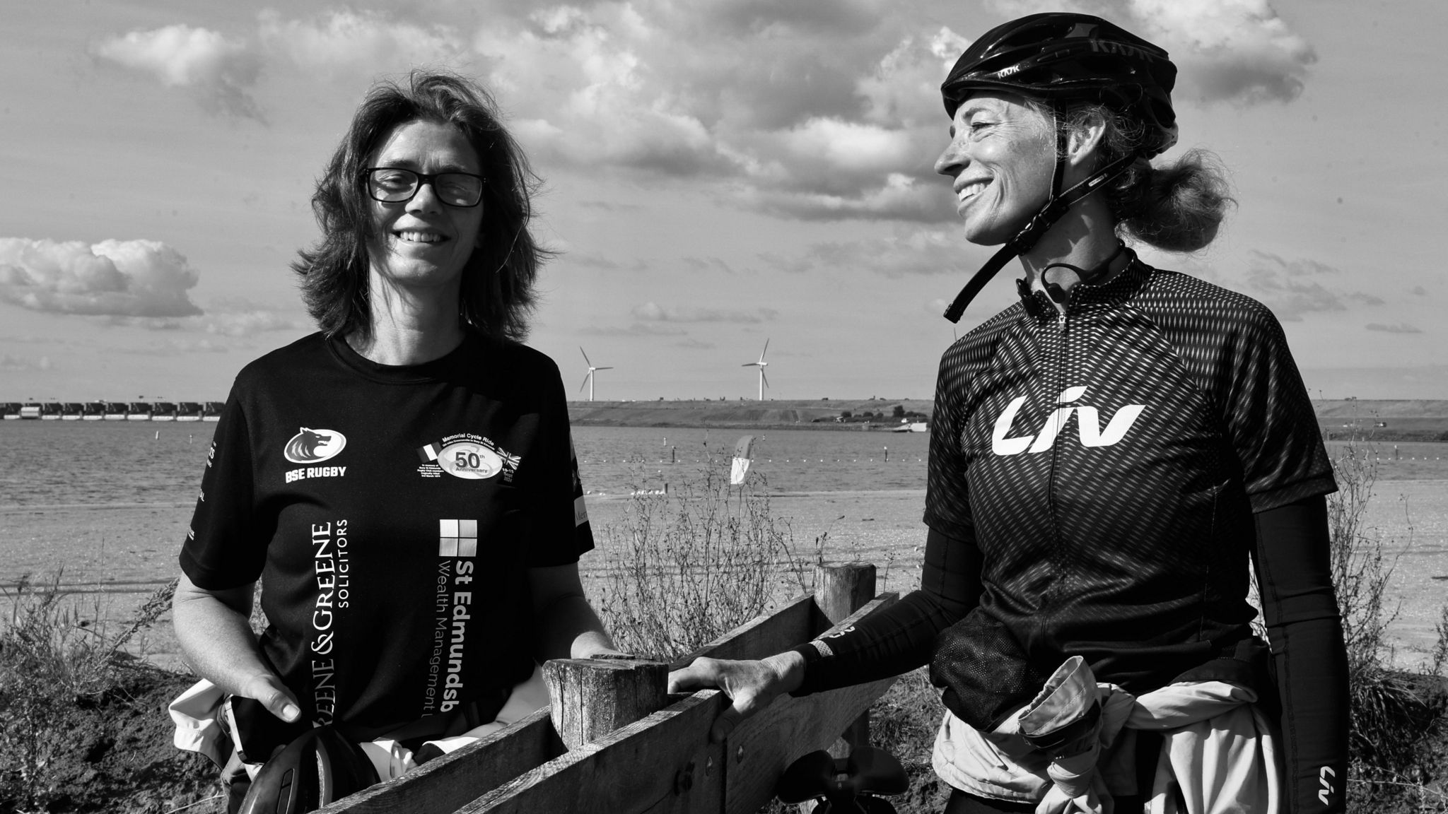 Black and white photo of two female cyclists. One is wearing a cycle helmet and smiling while the other sports a T-shirt with rugby club logos.