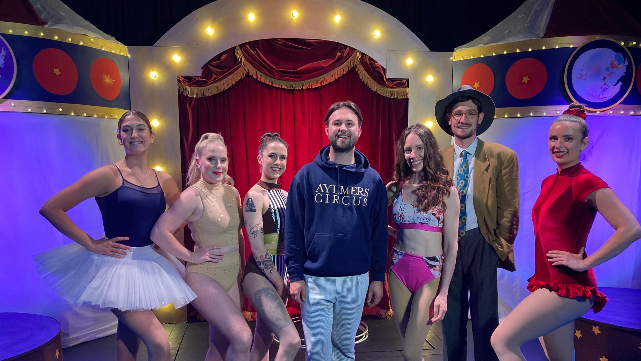 Harvey stands in the middle of a group of seven people. He's wearing a blue sweatshirt with three performers either side of him