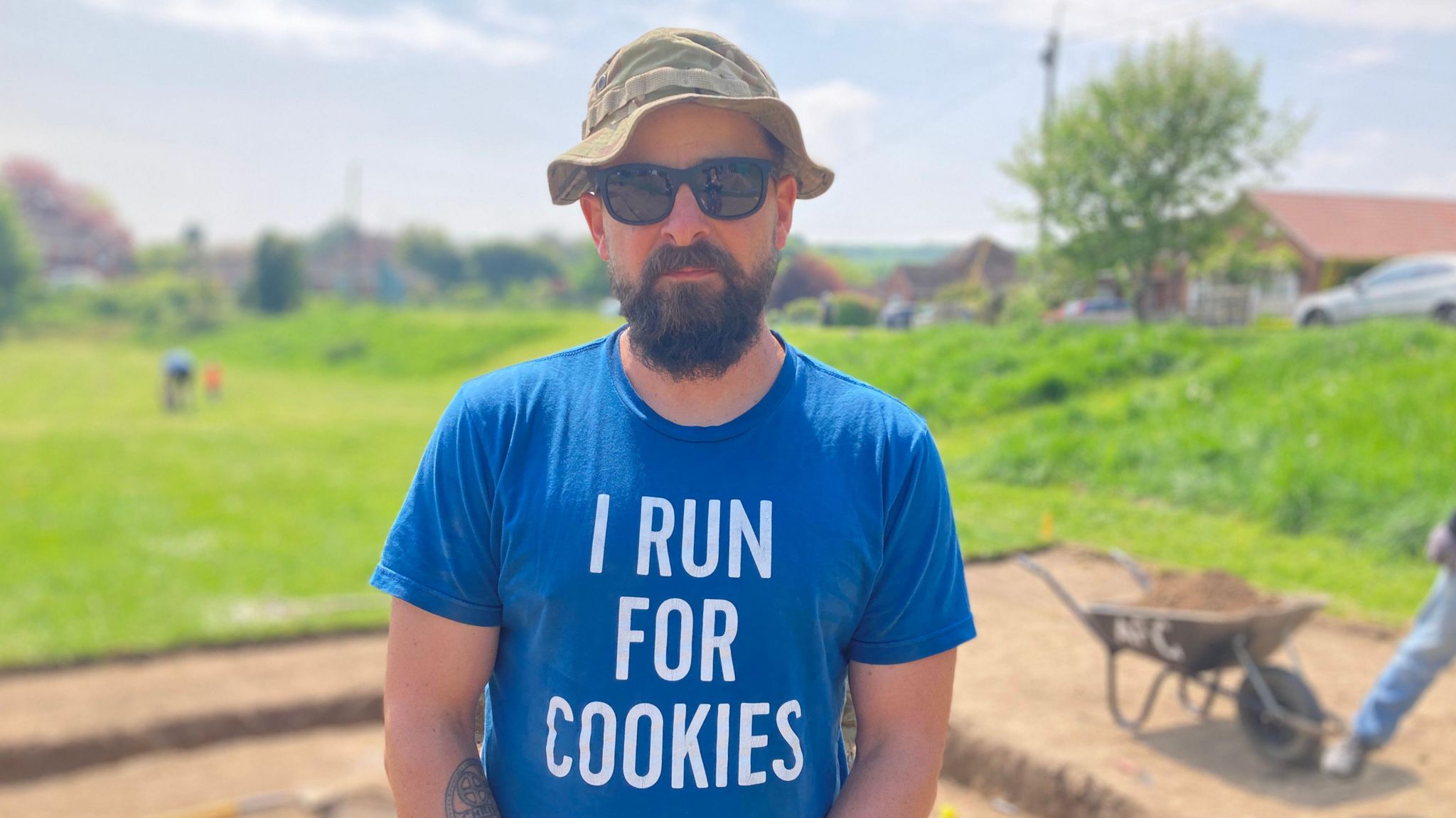 Patrick Carter looks at the camera from an archaeological dig site, wearing sunglasses, hat and a t-shirt saying 'I run for cookies'