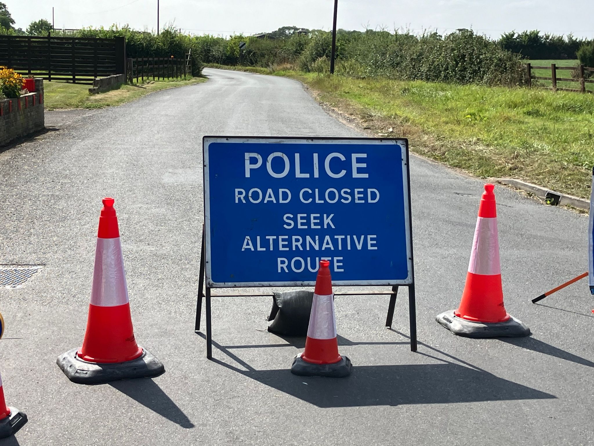 Blue police sign reads 'Police Road Closed Seek Alternative Route'. Sign is on country road and surrounded by traffic cones.