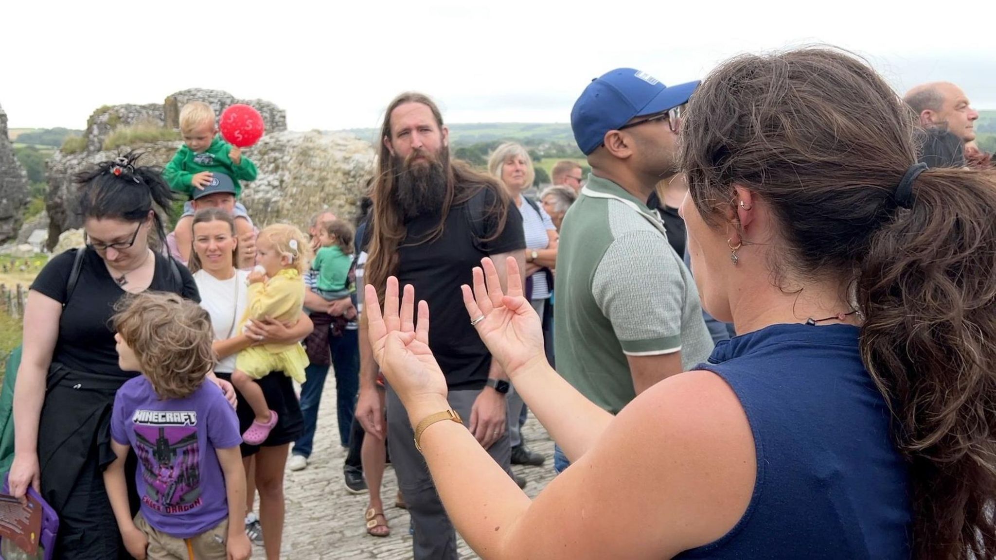 The BSL interpreter in front of a crowd of castle visitors. Some are looking at her interpretation while others are looking in the distance. It is a cloudy day.