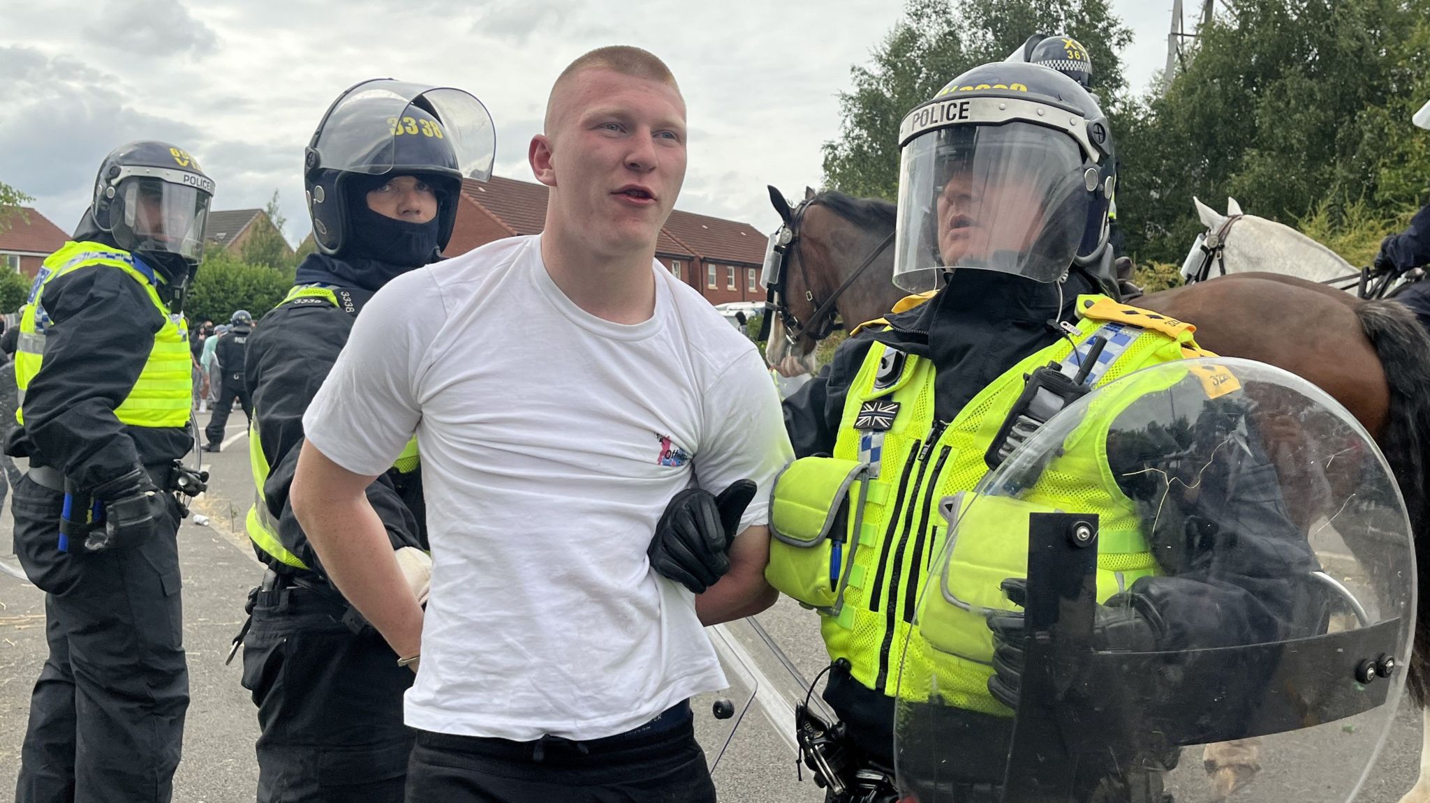 A man in a white t-shirt is led away by a police officer in riot gear