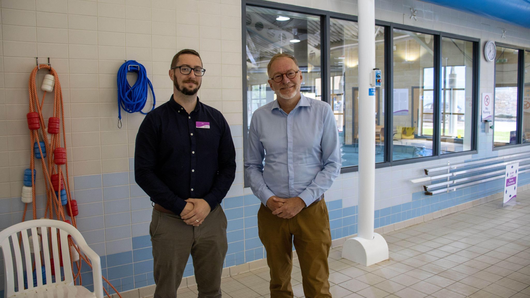 Two men looking at the camera, with their head down to their knees in view and standing near a swimming pool, which is out of vision. Both men wear glasses and sport a beard. The man on the left is wearing a dark blue shirt and grey-brown trousers. The man on the right is wearing a light blue shirt and brown trousers