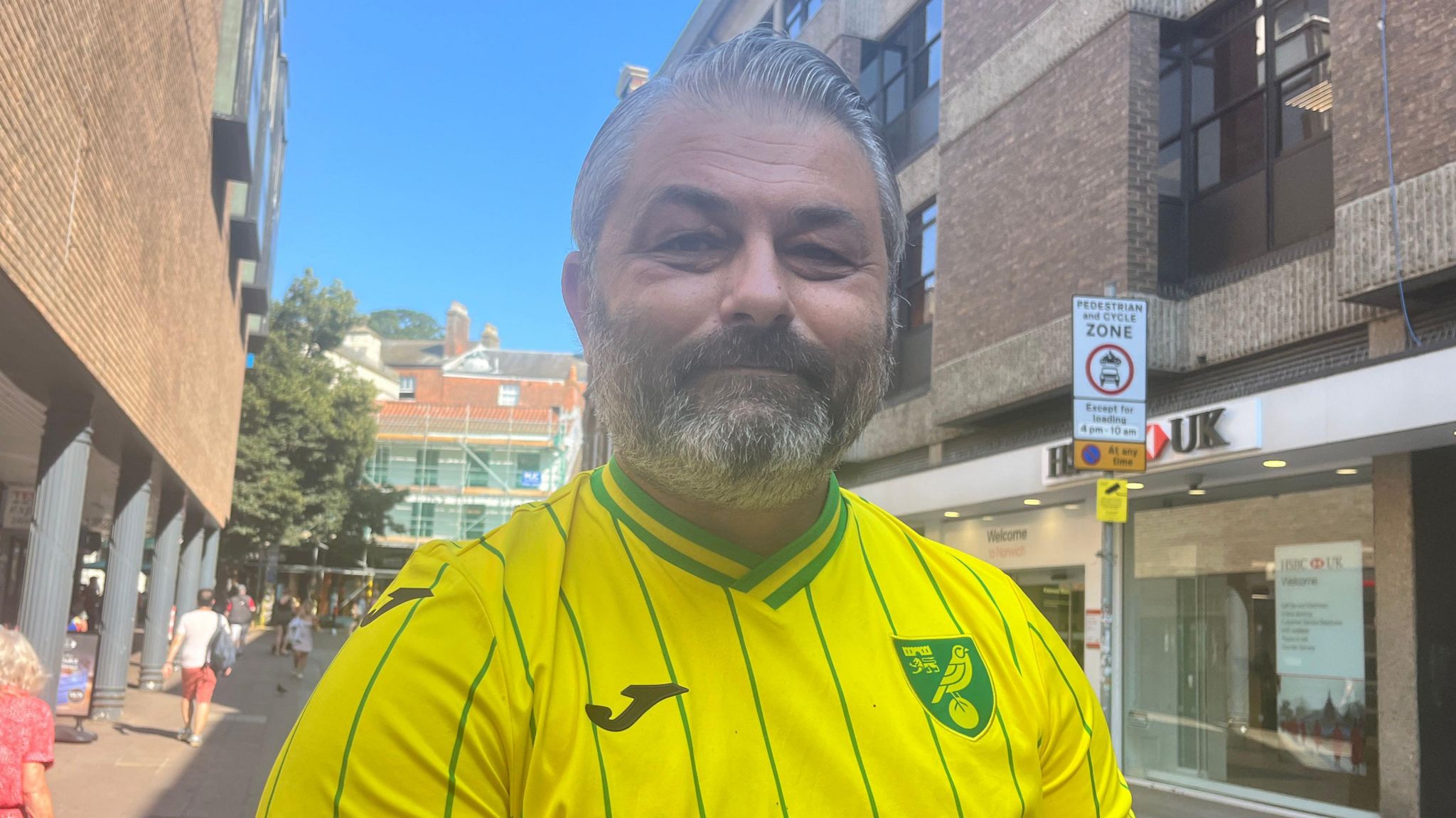 Simon Badderly smiles directly at the camera, wearing a yellow Norwich City football shirt. He is standing in a street. 