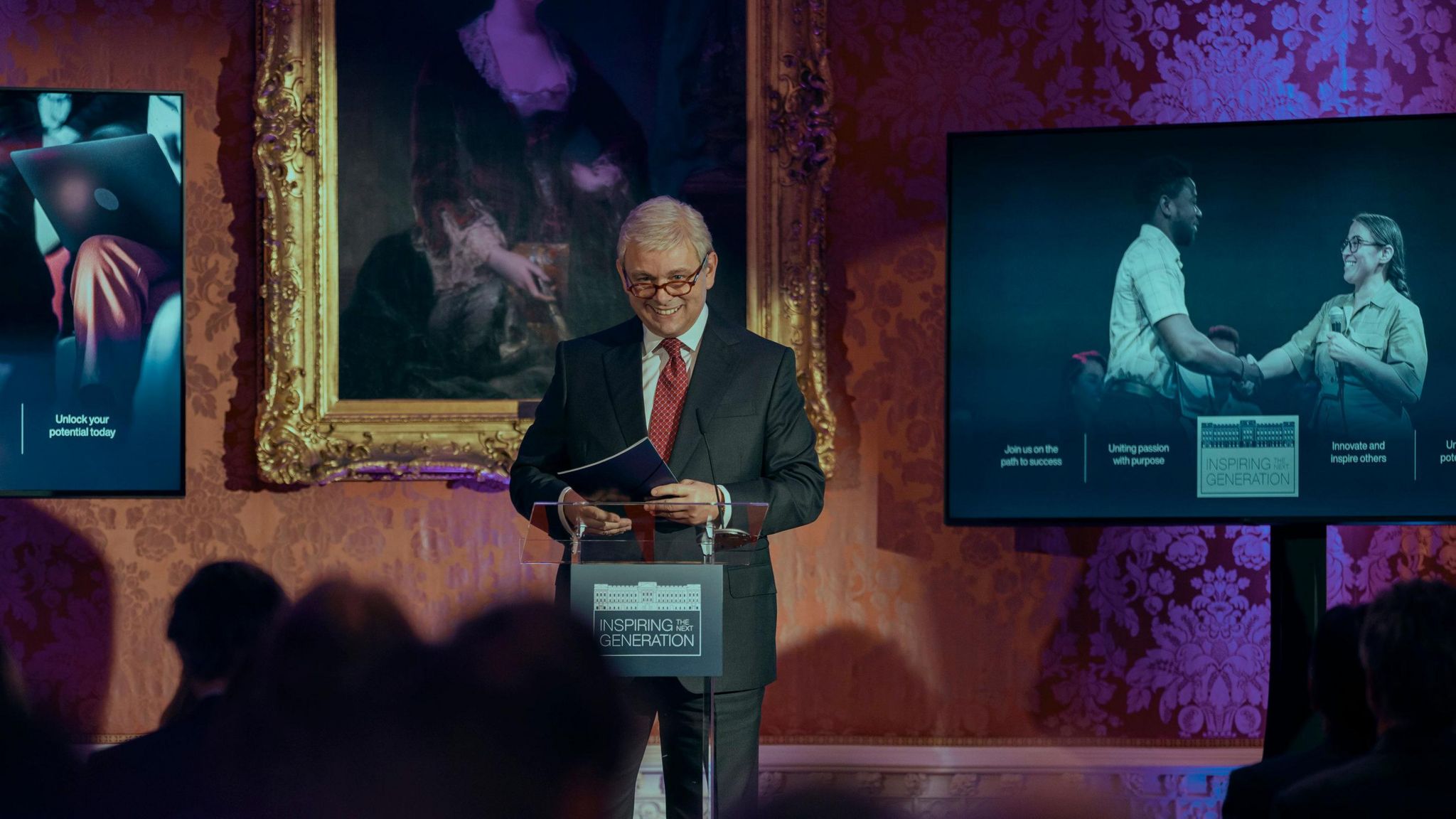 Michael Sheen, portraying Prince Andrew stands at a podium, smiling as he addresses a crowd of people, in front of posters and a portrait.