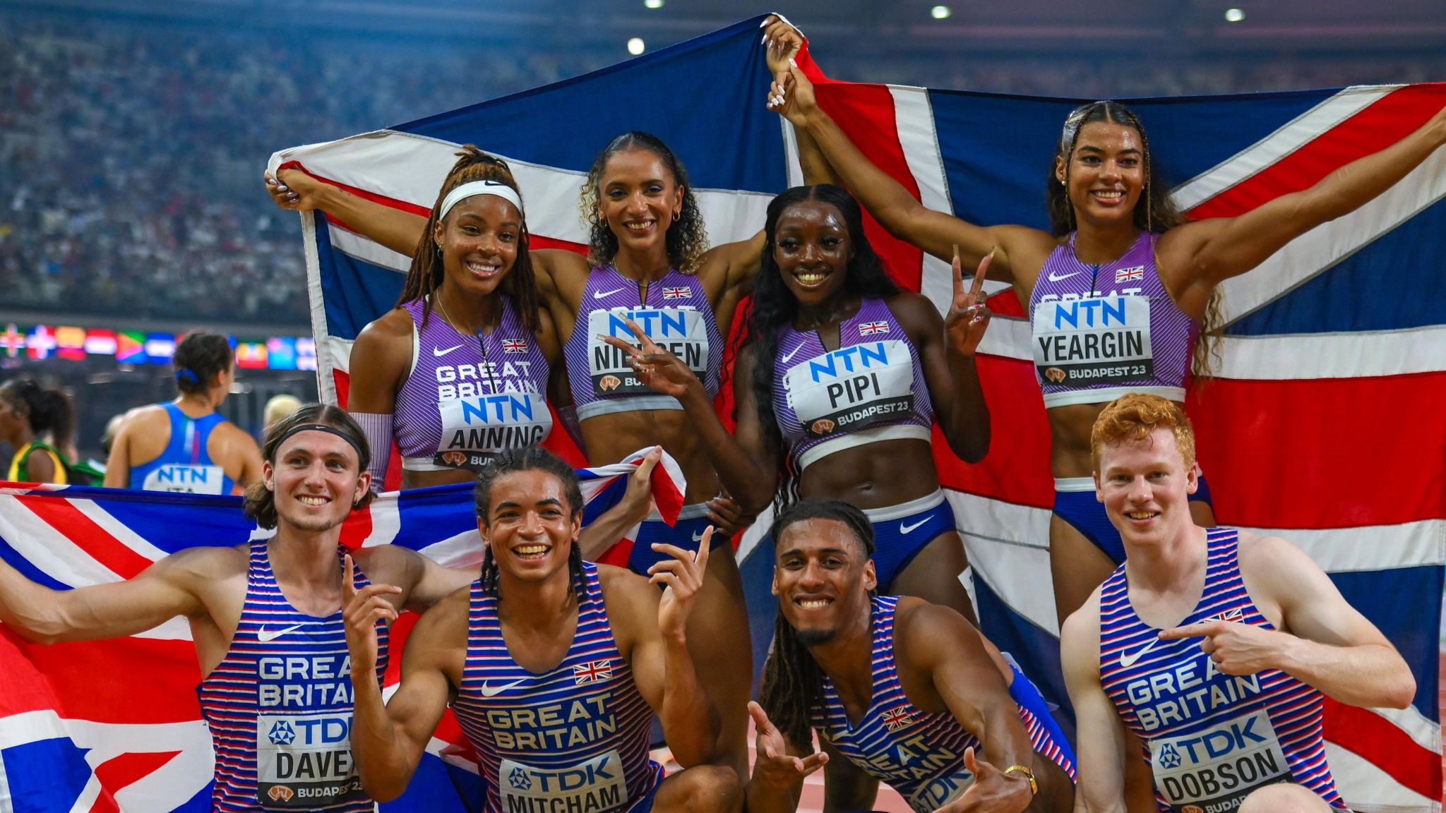 Four men are crouched down smiling at the camera with four women stood directly behind them also smiling at the camera. They are all dressed in red, white and blue running vests holding union jack flags