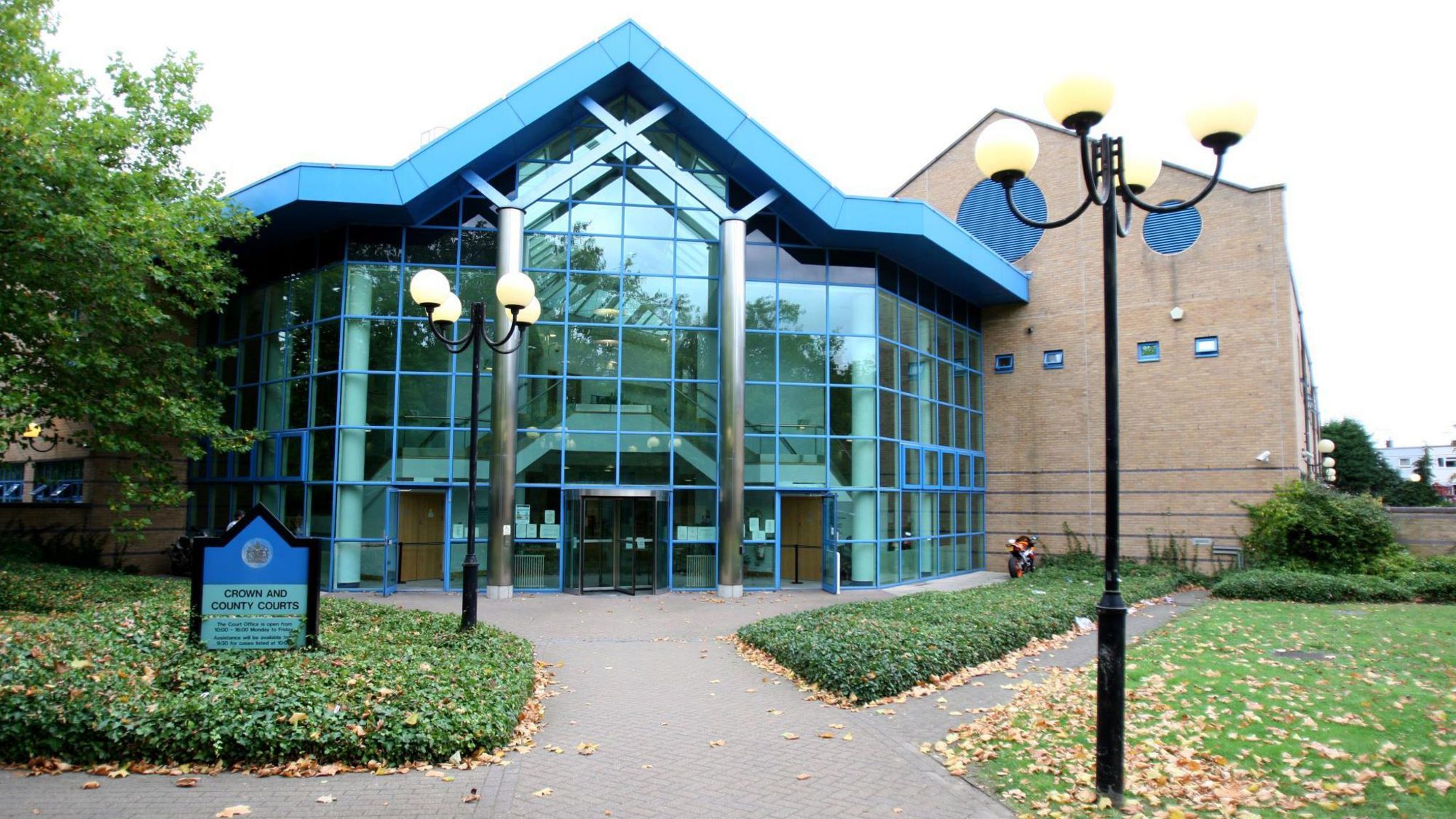 A glass-fronted building with window panes that have a blue trim. Outside is a grass area with a sign reading "Crown and County Courts". 