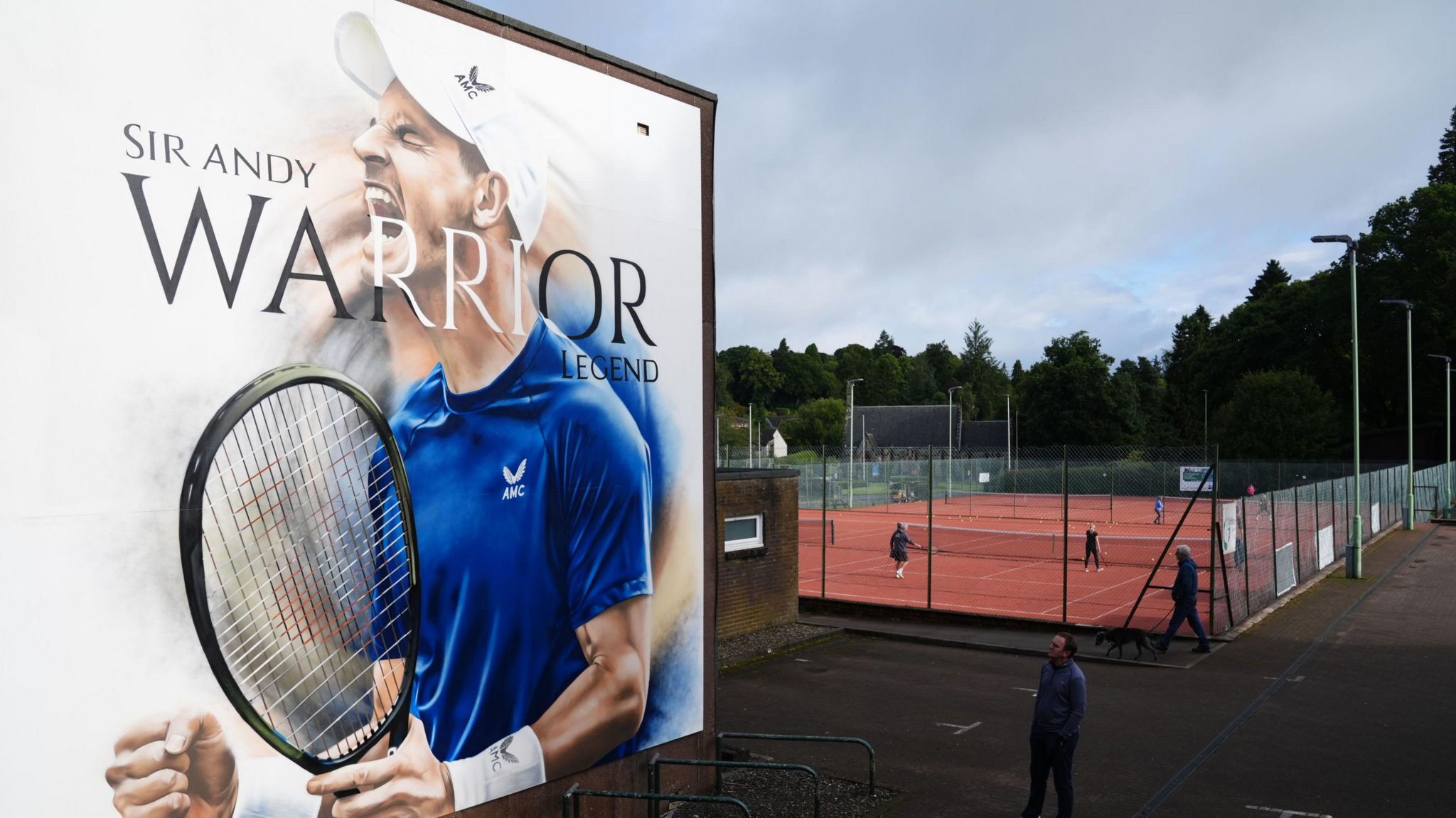 A 20ft mural of Andy Murray holding a tennis racket with his hands grasped and mouth opens. It reads Sir Andy, warrior, legend.