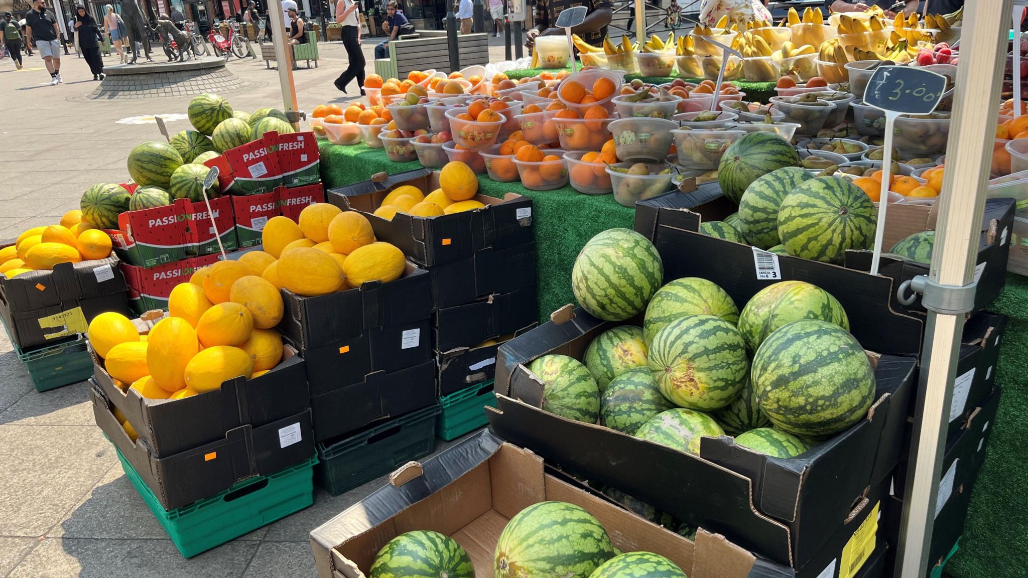 A fruit stall in Uxbridge displaying large boxes containing watermelons and yellow honeydew melons along the pavement. On the table there are several bowls containing oranges, pears and bananas.