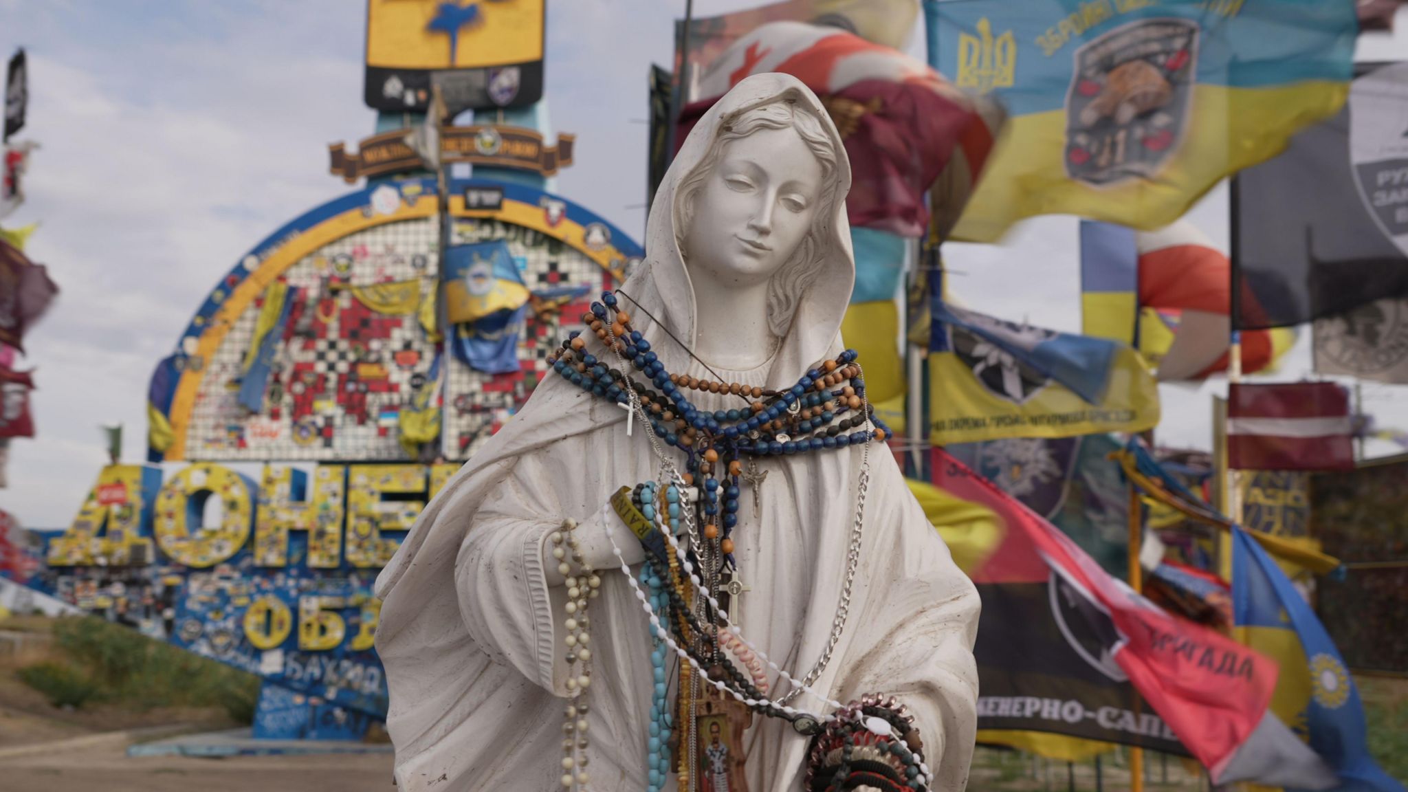 A statue of a woman is draped in rosary bead necklaces, with Ukrainian unit brigade flags draped on road signs behind it