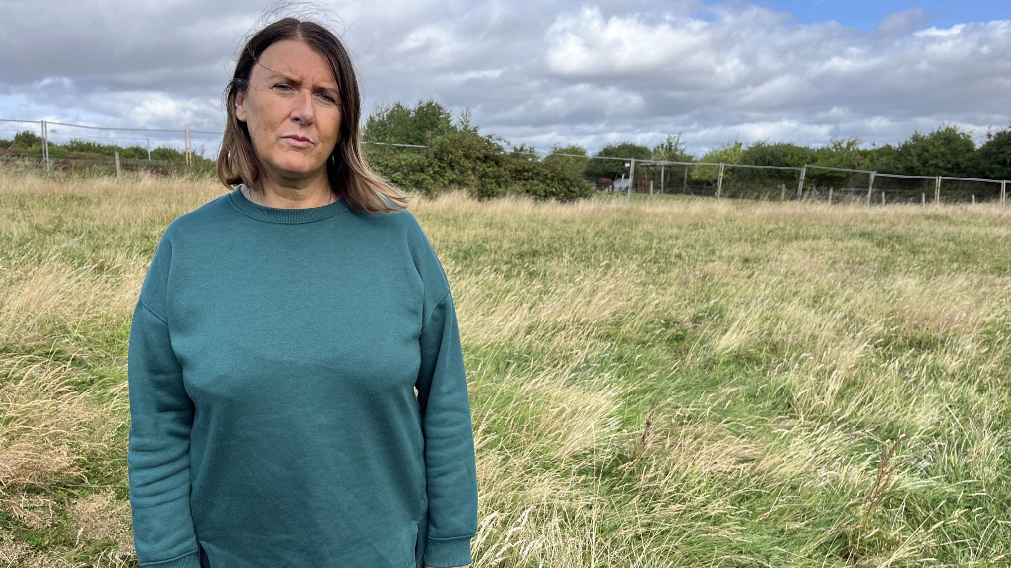 A woman with brown hair standing in a green field. She is wearing a green- blue-coloured top.