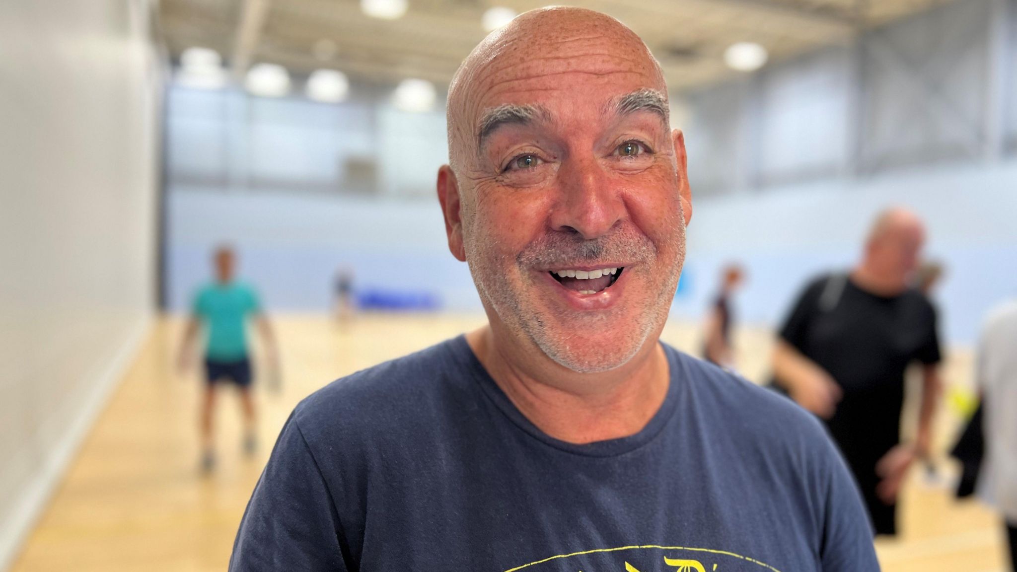 Rab McEwan is wearing a navy t-shirt. He has a bald head and facial hair. He is smiling at the camera. In the blurred background, you can see a sports hall with blue walls and a wooden floor