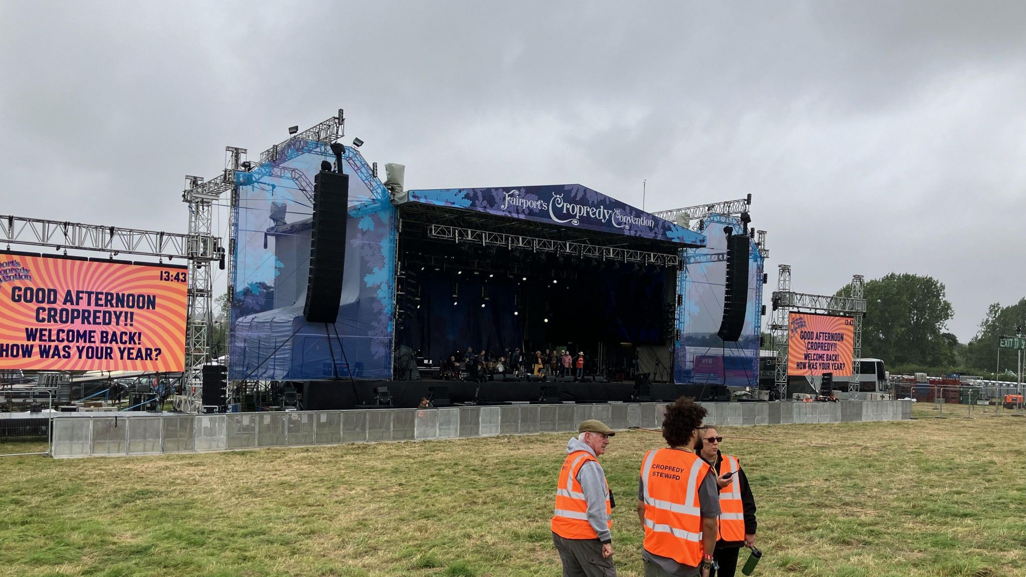 A large stage with Fairport's Cropredy Convention written at the top. There is a green field with people wearing hi-vis jackets in front of the stage