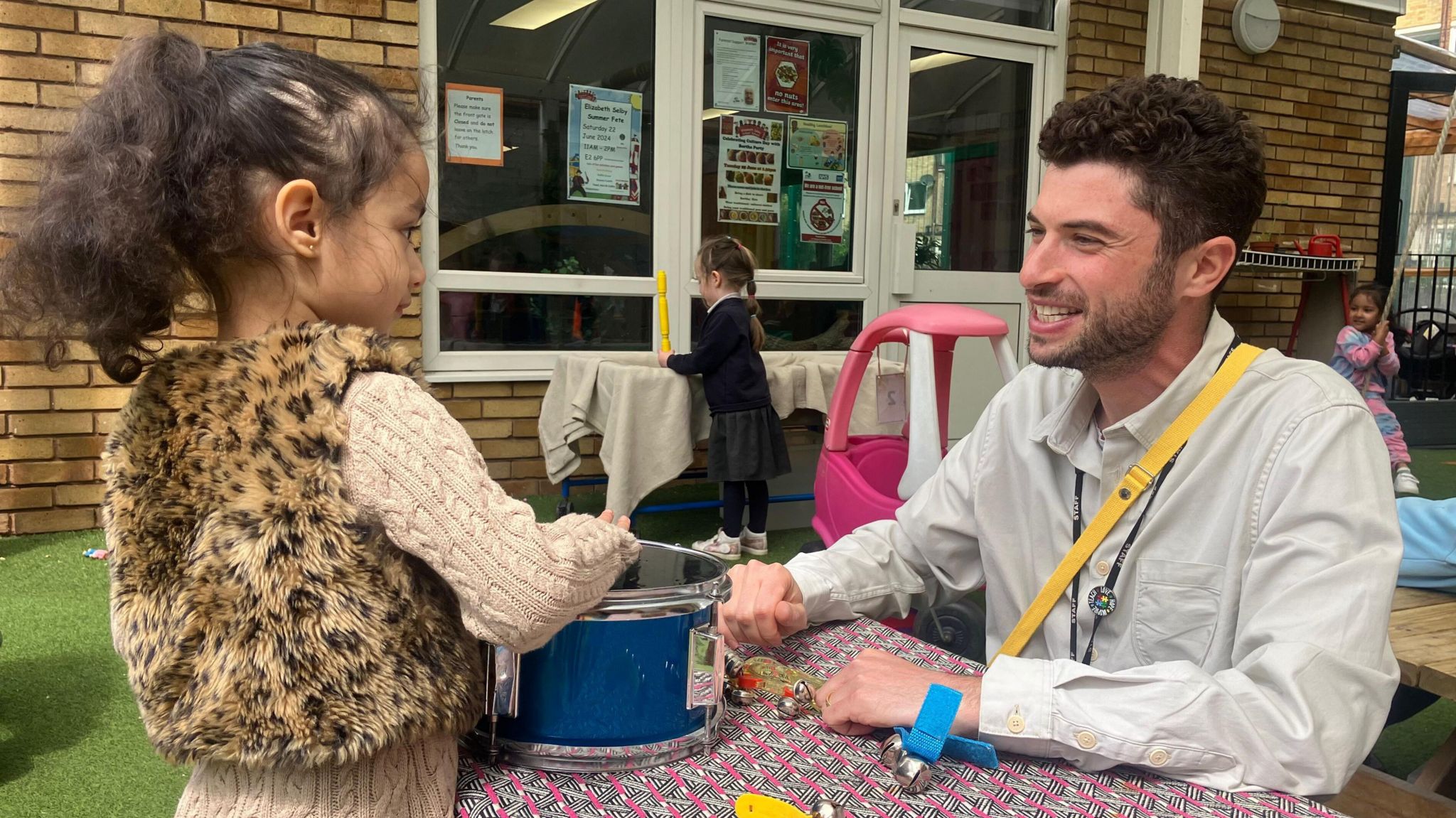 A man with brown hair and a beige shirt sits at a table smiling at a young girl with curly hair and a leopard body warmer and beige woollen cardigan. She is playing a blue drum and smiling back. 