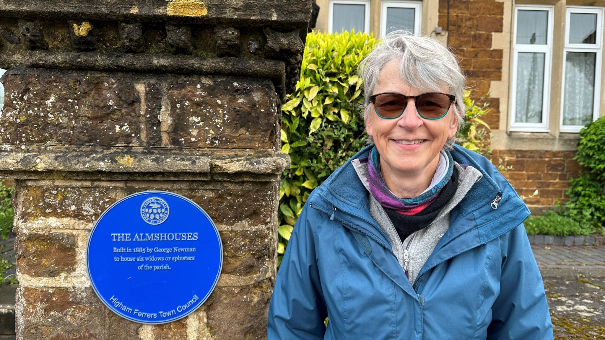 Cathy Kennedy standing by a blue plaque in Higham Ferrers, Northamptonshire