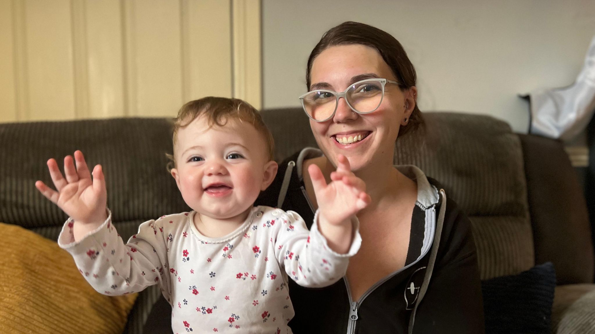 Women in black hoodie and wearing glasses smiling at the camera. She is sat in a living room on a sofa. She has a baby girl sat on her lap with her siling and putting her hands up in the air.