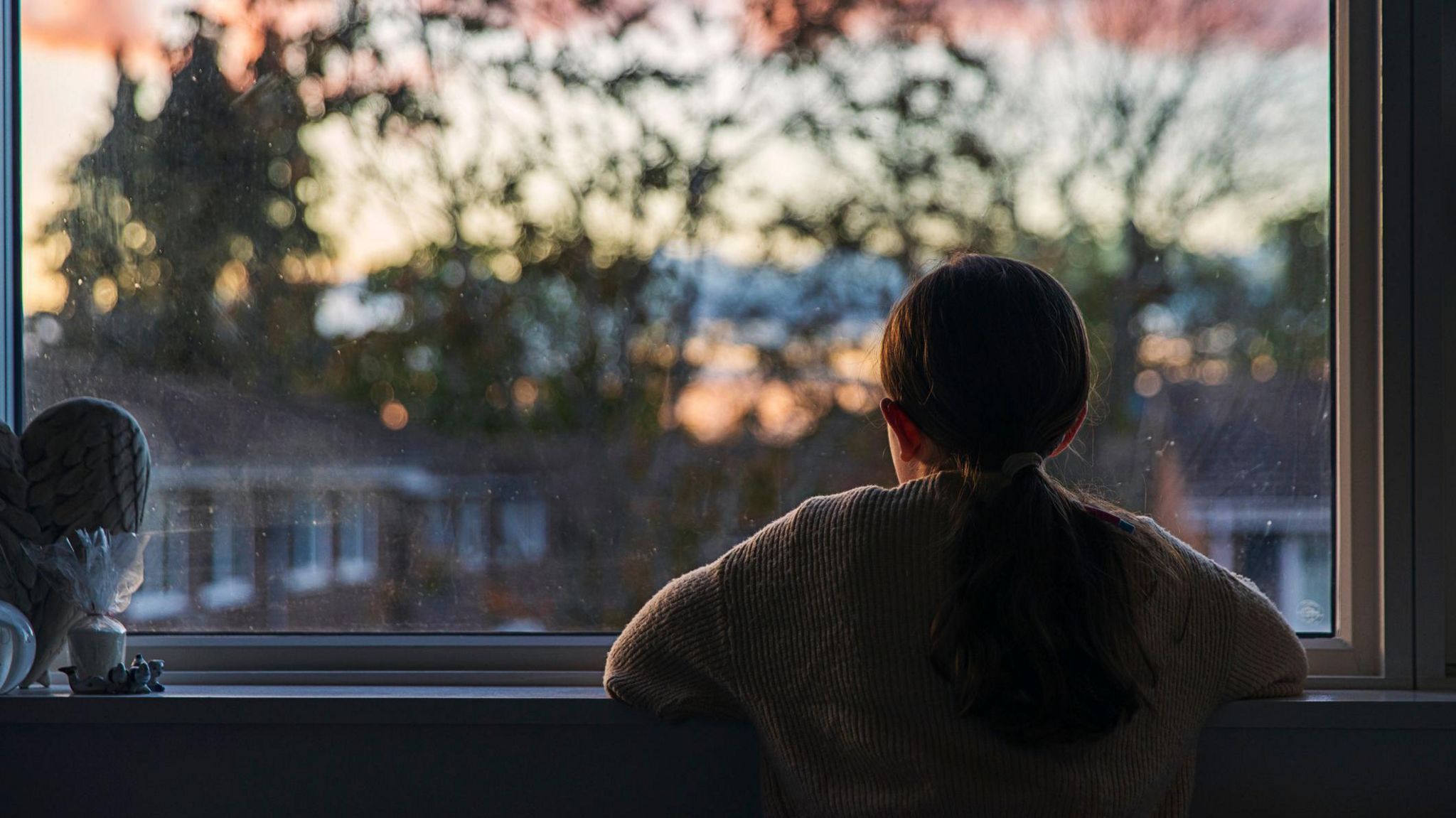 A young girl looks out of a window at a darkening sky. Seen from behind, she is wearing a jumper and has her hair in a ponytail