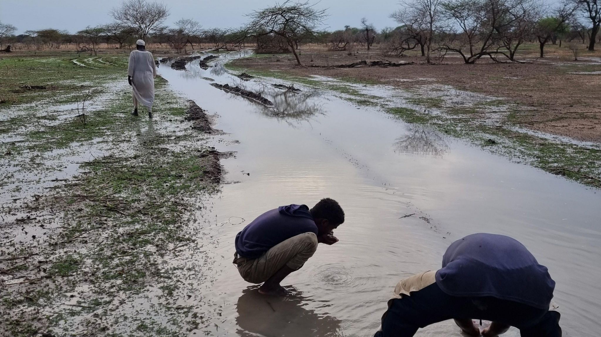 Two men crouch over a pool of rainwater in the road as the travel out of Sudan