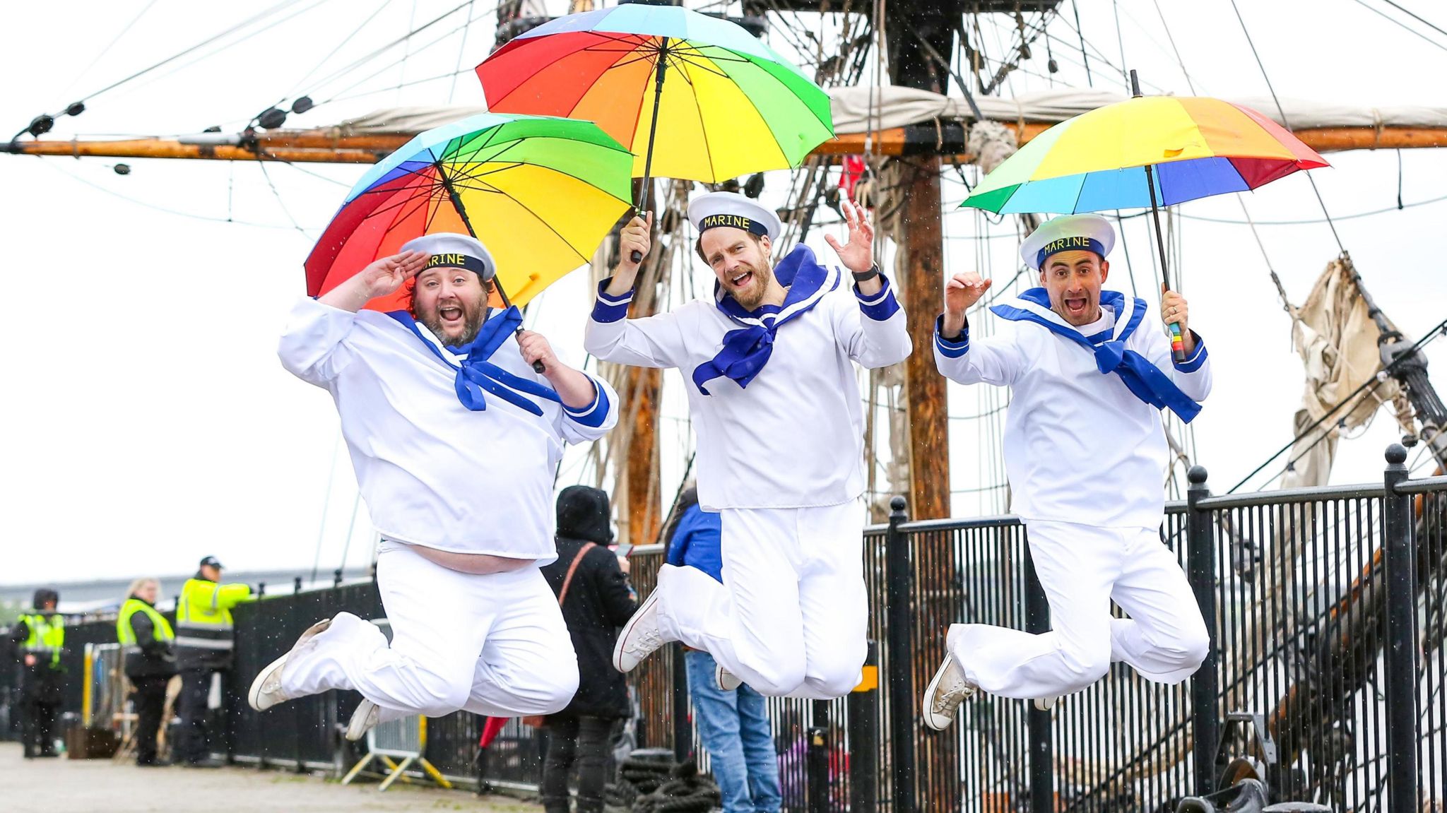 Three men stood in front of a tall ship and dressed as sailor jump in the air while holdiing multi coloured umbrellas