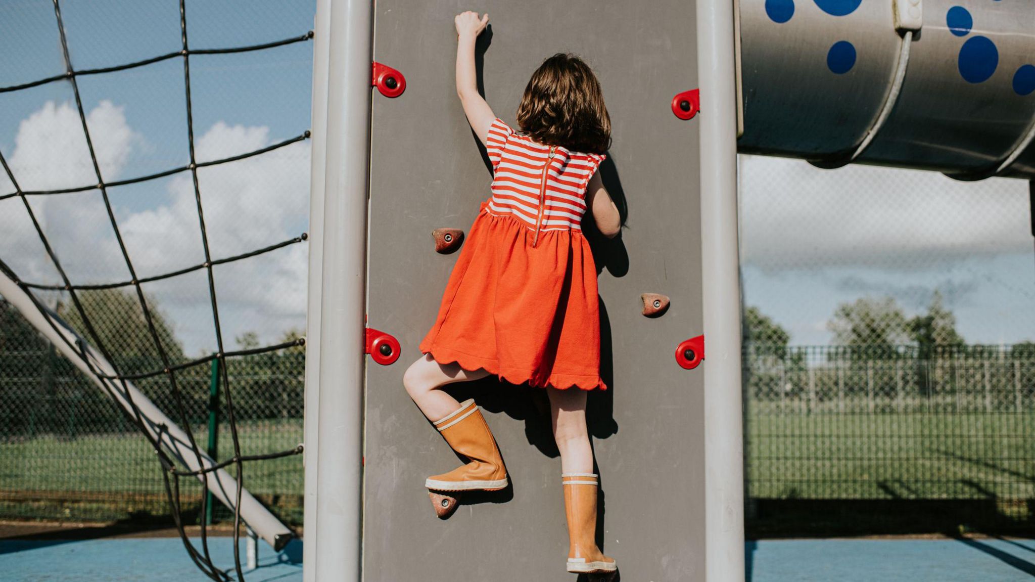 A little girl scales a climbing wall in a play park on a sunny day.