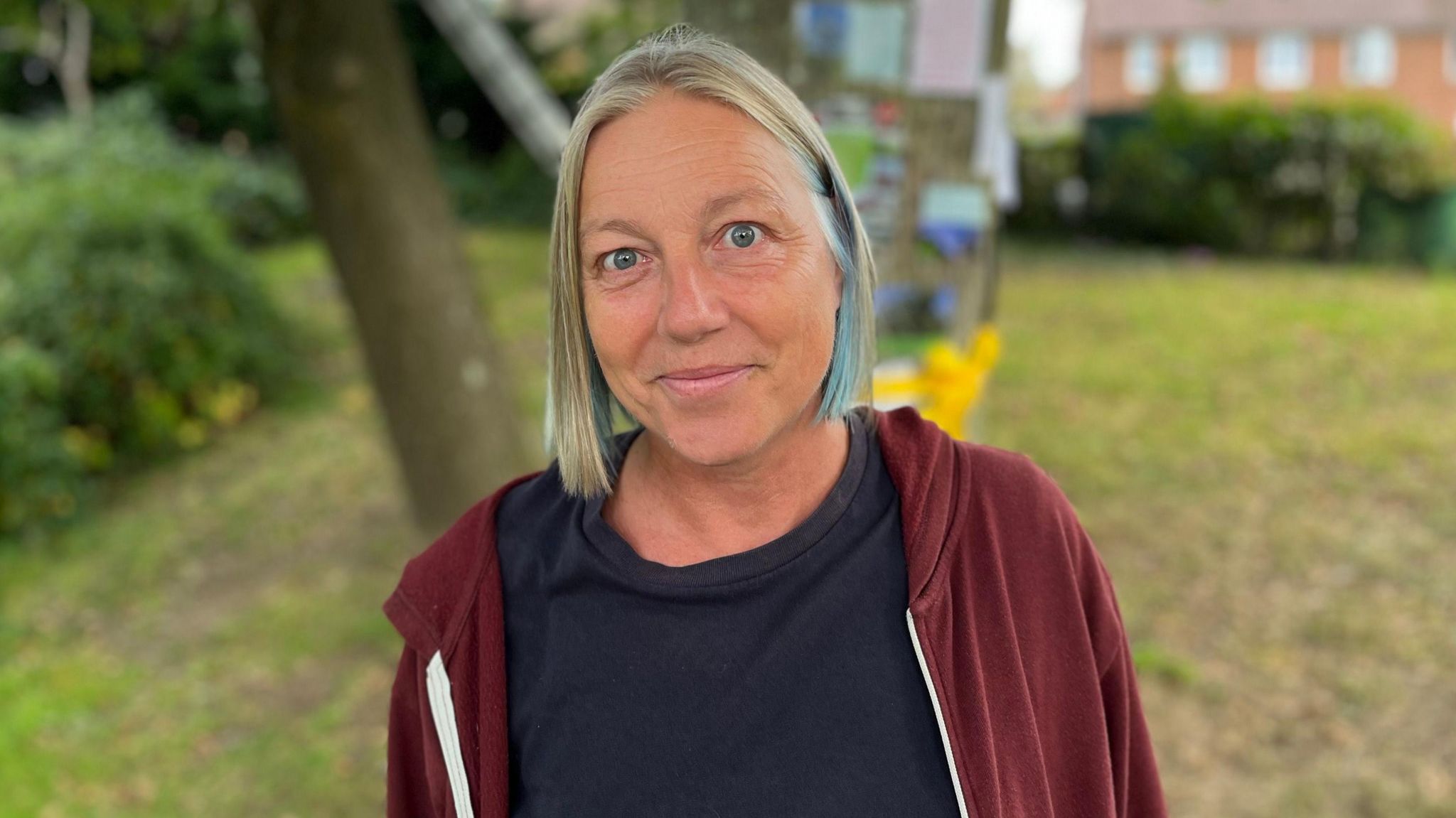 Main organiser Gabi Barrett stands in front of the oak tree and smiles at the camera. She has fair bobbed hair with a blue streak in it and wears a dark red hoodie and a black t-shirt