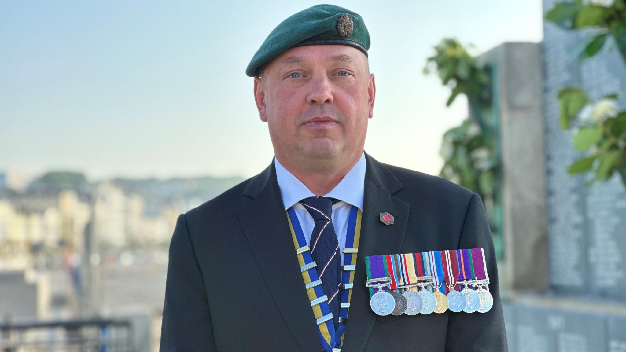 A man wearing a green beret with medals on his chest with a war memorial behind him