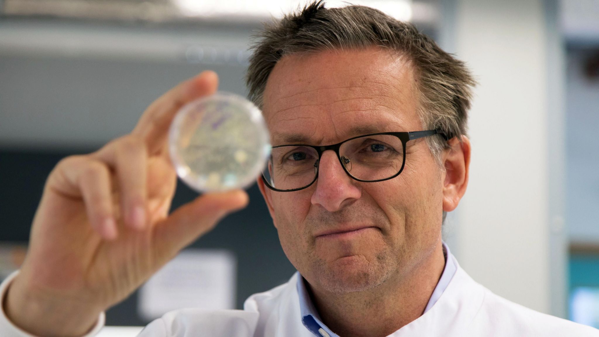 Dr Michael Mosley, wearing a white lab coat and blue shirt, holds up a translucent ball to the camera