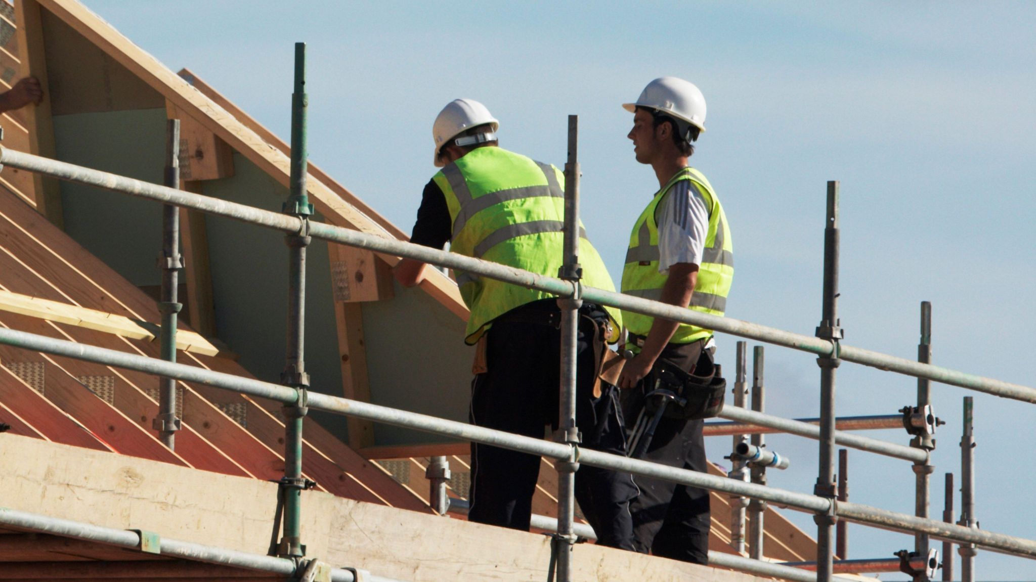 Two builders wearing hi-vis yellow vests and hard hats. They are standing on scaffolding tending to the roof of a building