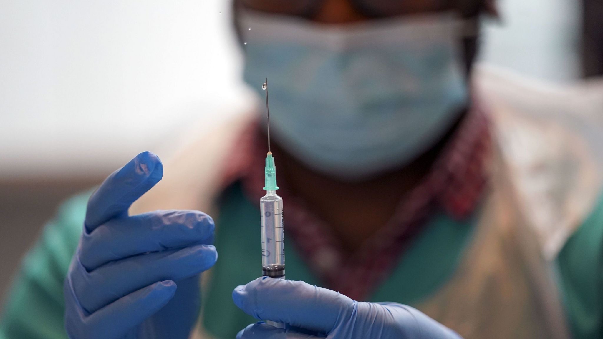 A medical professional in protective clothing and a mask prepares a syringe.