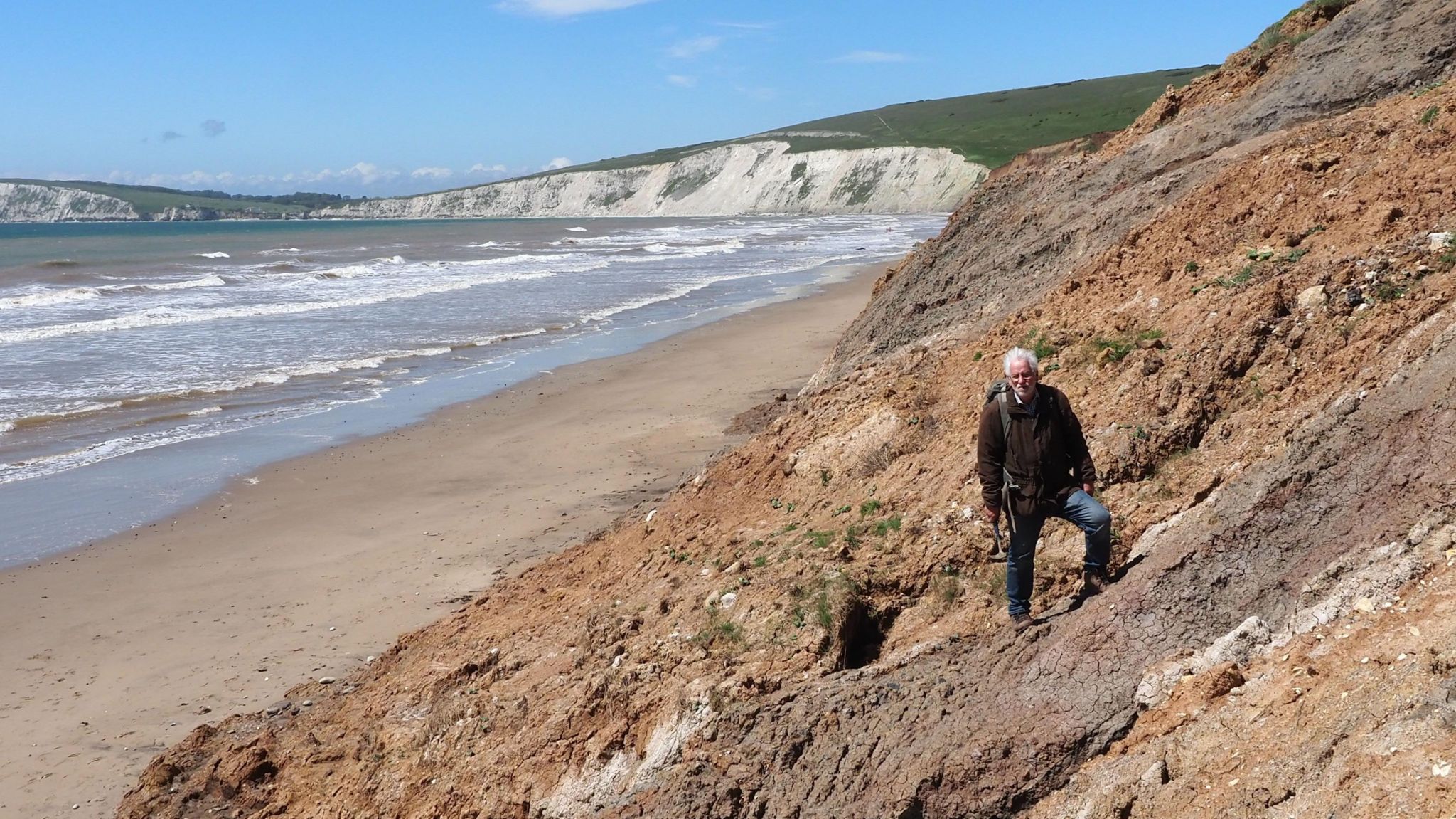 Jeremy Lockwood standing on crumbling sloped cliffs. He has white hair and is wearing a large coat and jeans. Behind him is the beach and sea and white cliffs in the distance