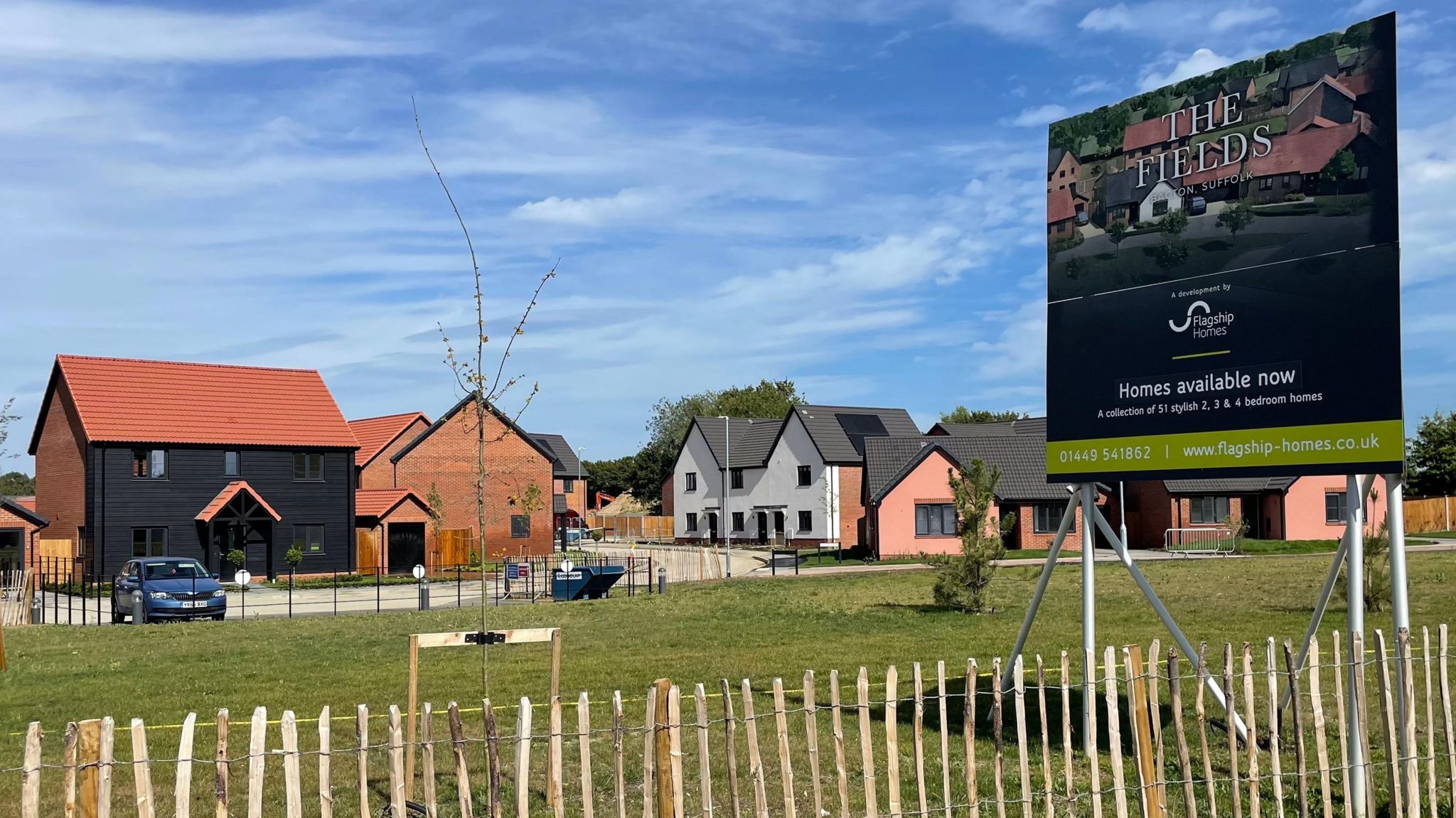 New houses on The Fields development. Around five houses can be seen in the middle distance, made of a range of materials. One is black clapperboard, another is white and others are red brick. Their roofs are either grey tile or red tile. A low-level wooden fence runs along the bottom of the picture, with grass and young trees in the foreground. A billboard on the right advertises The Fields and says "homes available now".