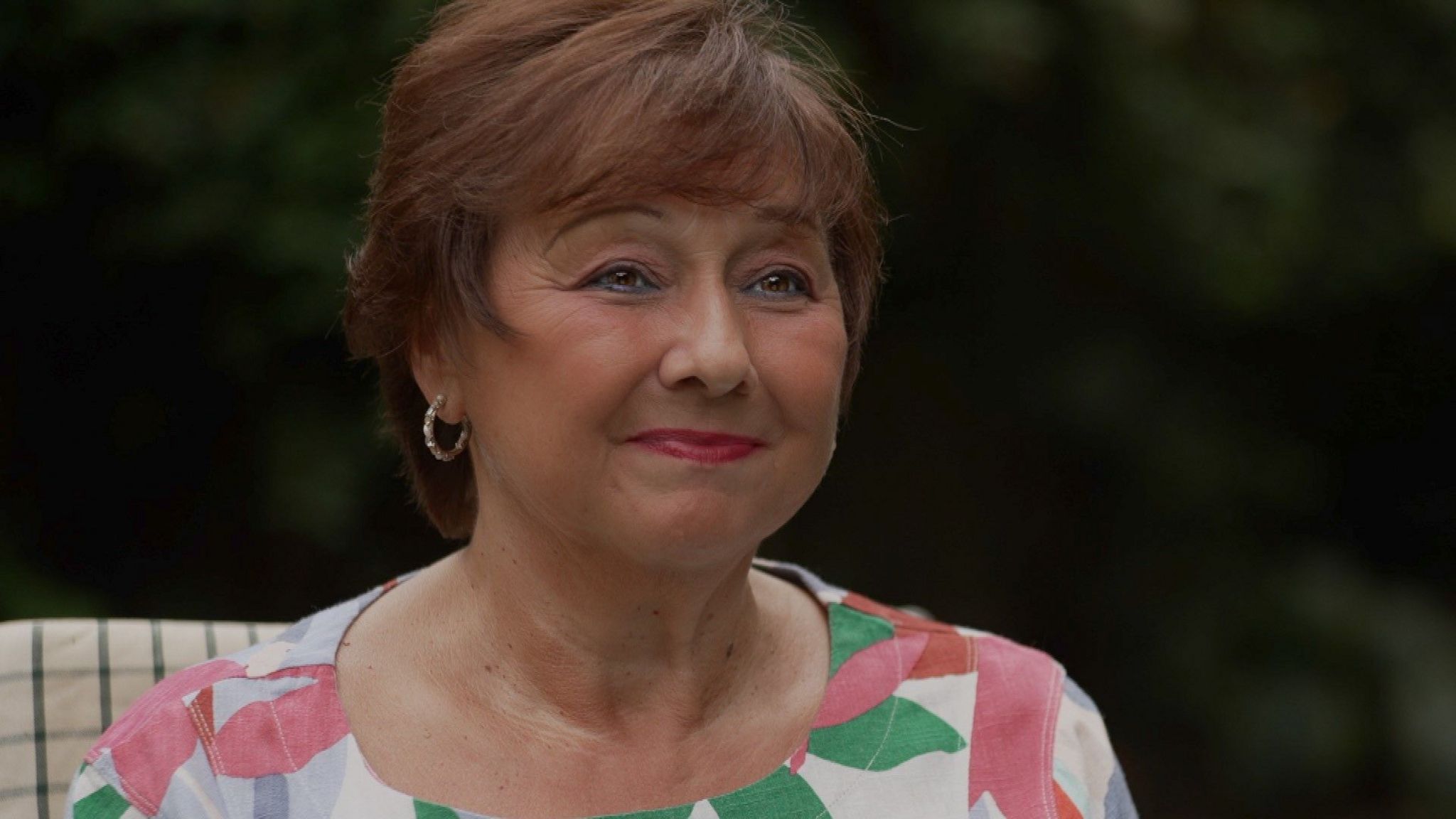 Guiliana Rossi sitting down on a chair, and looking towards the right, she is wearing a pink and white floral top, has short cropped brown hair and has gold hoop earrings.