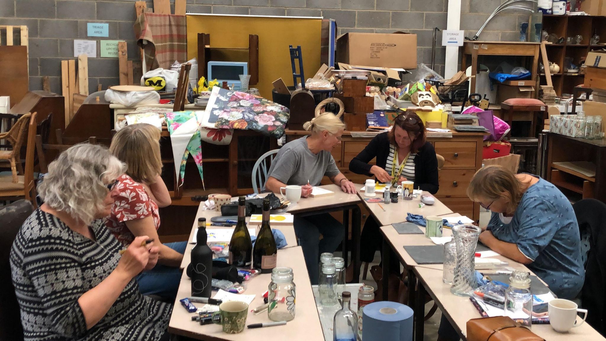 A group of five people are pictured around a table, with a variety of items including wine bottles, mugs, vases and paper which they are upcycling