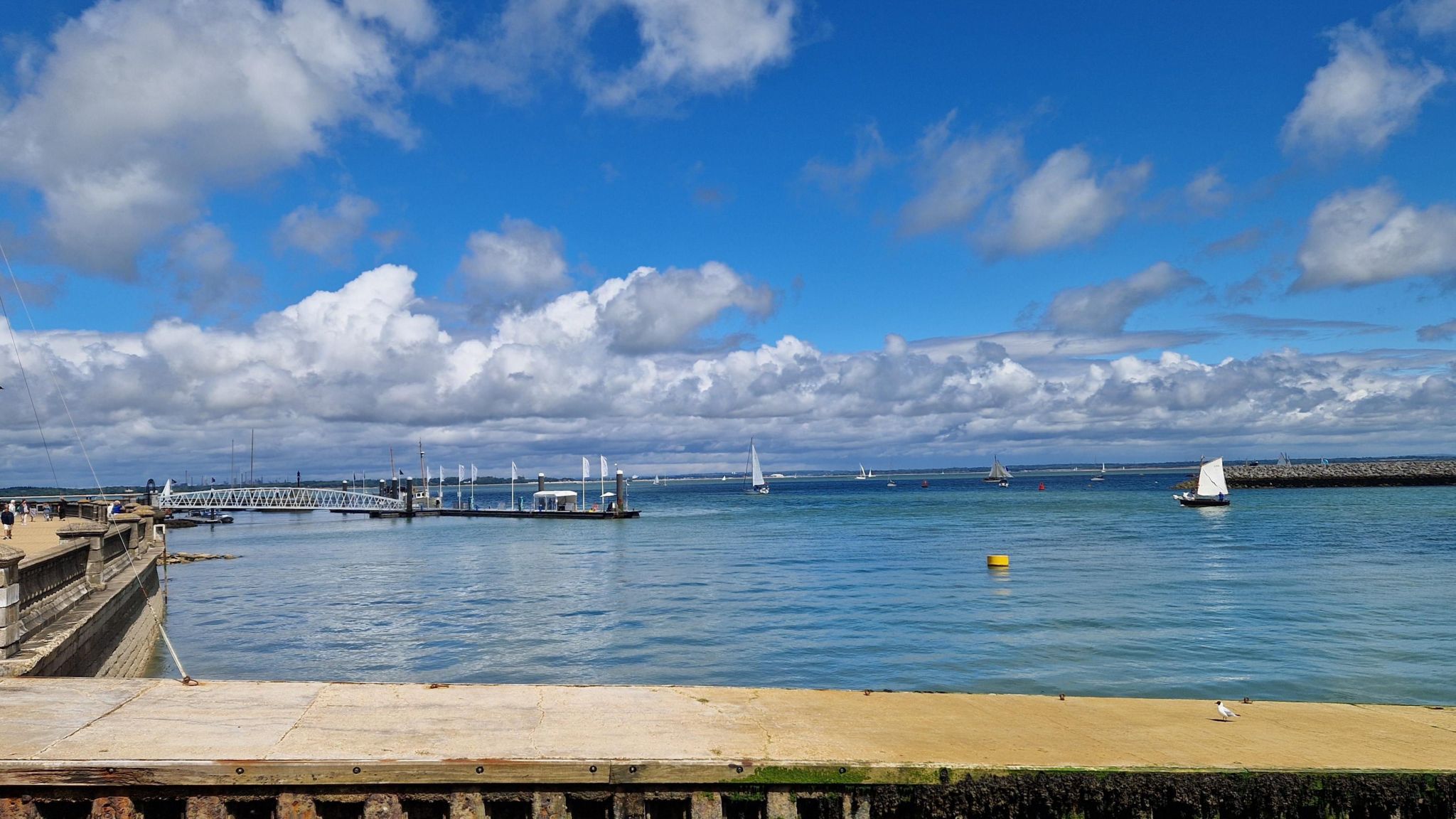 THURSDAY - Cowes seawall is in the foreground with the harbour behind. There are three sailing boats with white sails. There is a pontoon with a walkway and a yellow buoy in the water. Behind there is the breakwater and the blue water of the Solent with several more sailing boats. In the distance is the mainland. The sky above is blue with white clouds