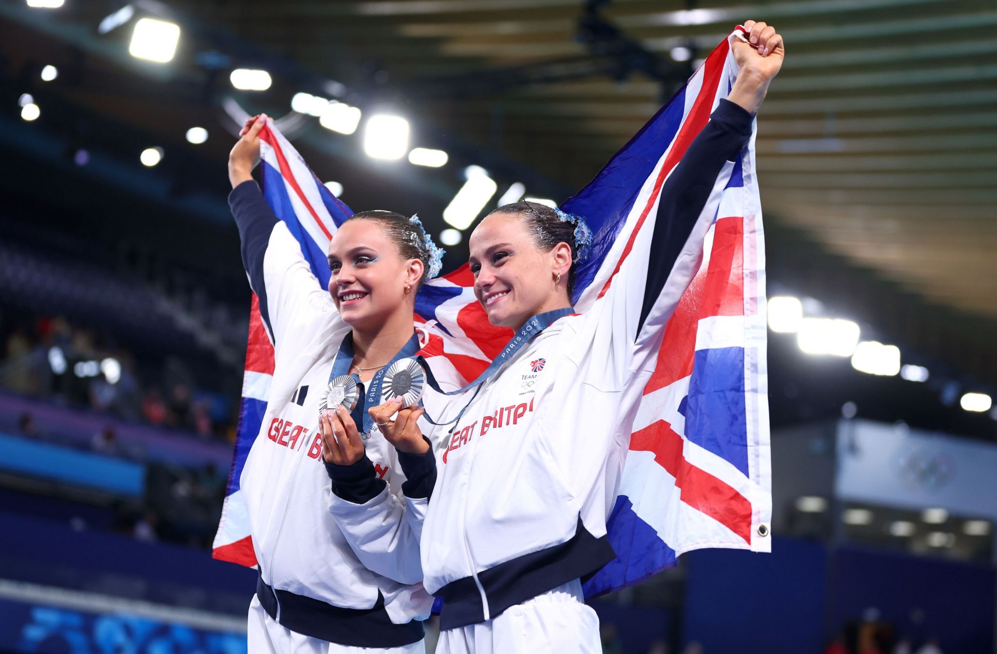 Kate Shortman and Izzy Thorpe stand smiling, holding up a Union Jack flag behind them. They are wearing white tracksuits and are holding their silver medals up.