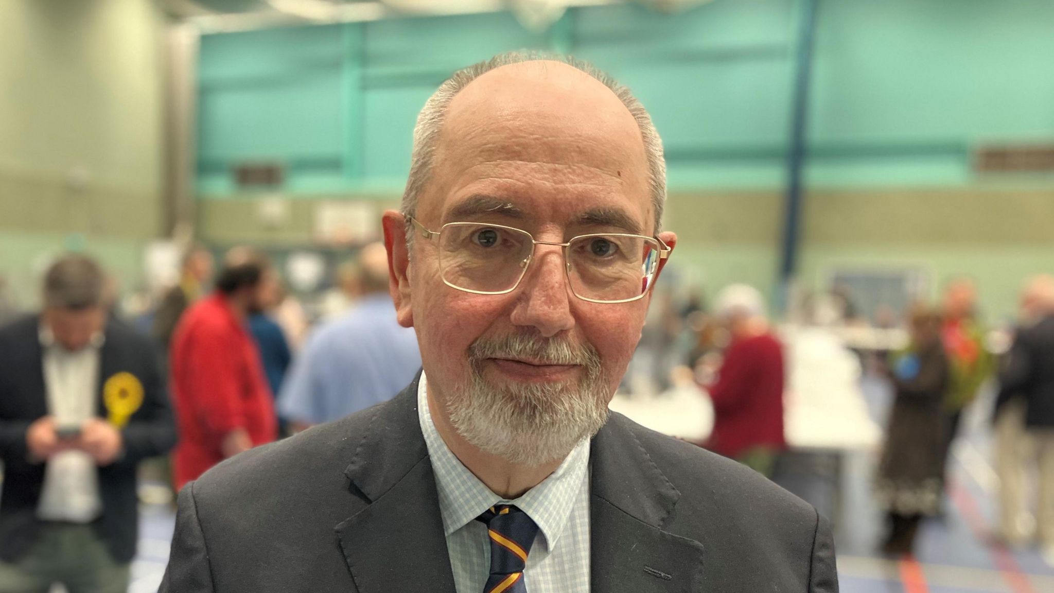 A bald man with glasses and a short grey beard, wearing a suit and tie photographed from the torso up. A sportshall is out of focus in the background.