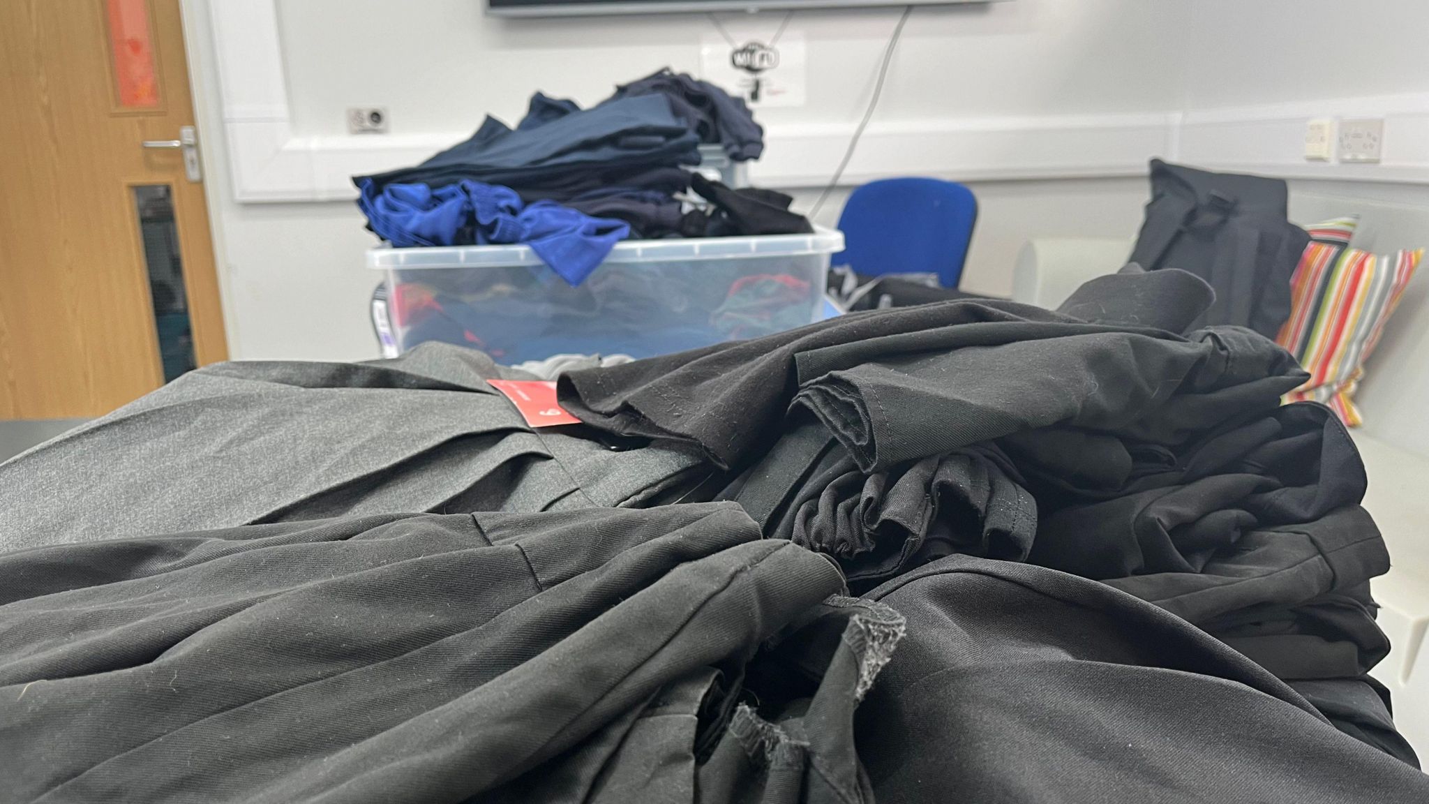 A pile of black pieces of school uniform clothing, with a clear plastic box in the background containing more.