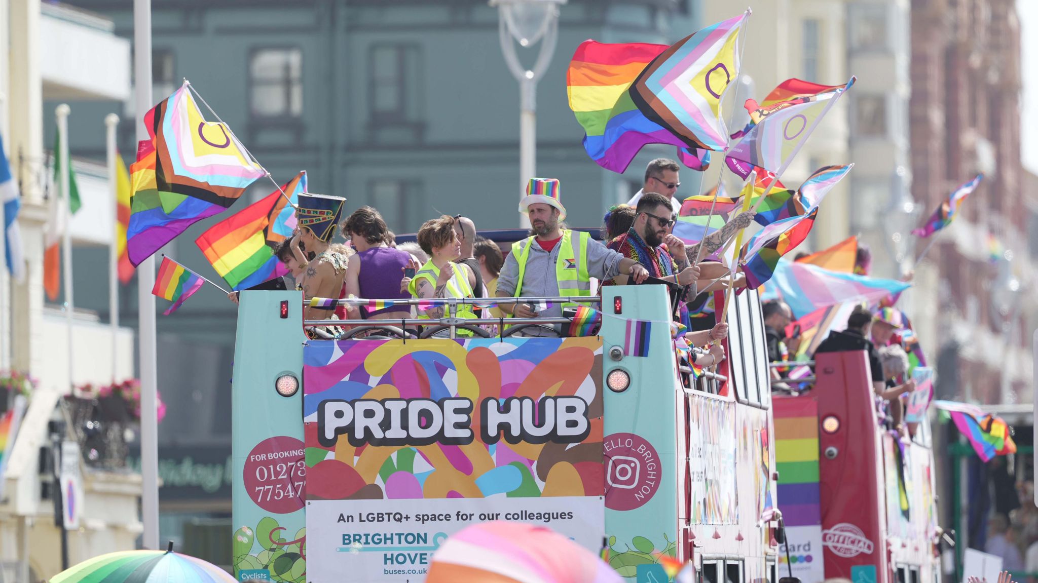 Pride Hub buses making their way along Kings Road. People are stood on the top deck of the open top buses waving rainbow flags