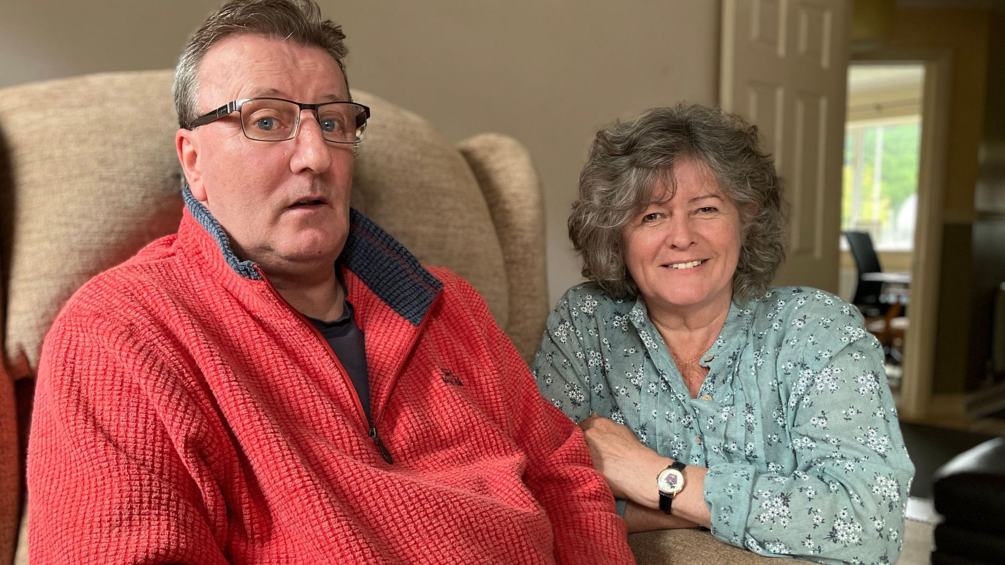 Rob Sizer sits on a dark brown leather sofa with his wife Ann, the wall behind them is red. Rob is on the left wearing frameless glasses and a red jumper with a zip and collar holding his hands in his lap, while Ann sits on the right, she is smiling looking slightly off camera while she talks to the reporter. Ann has short grey wavy hair and wears a pale green blouse with a white flower print on it.