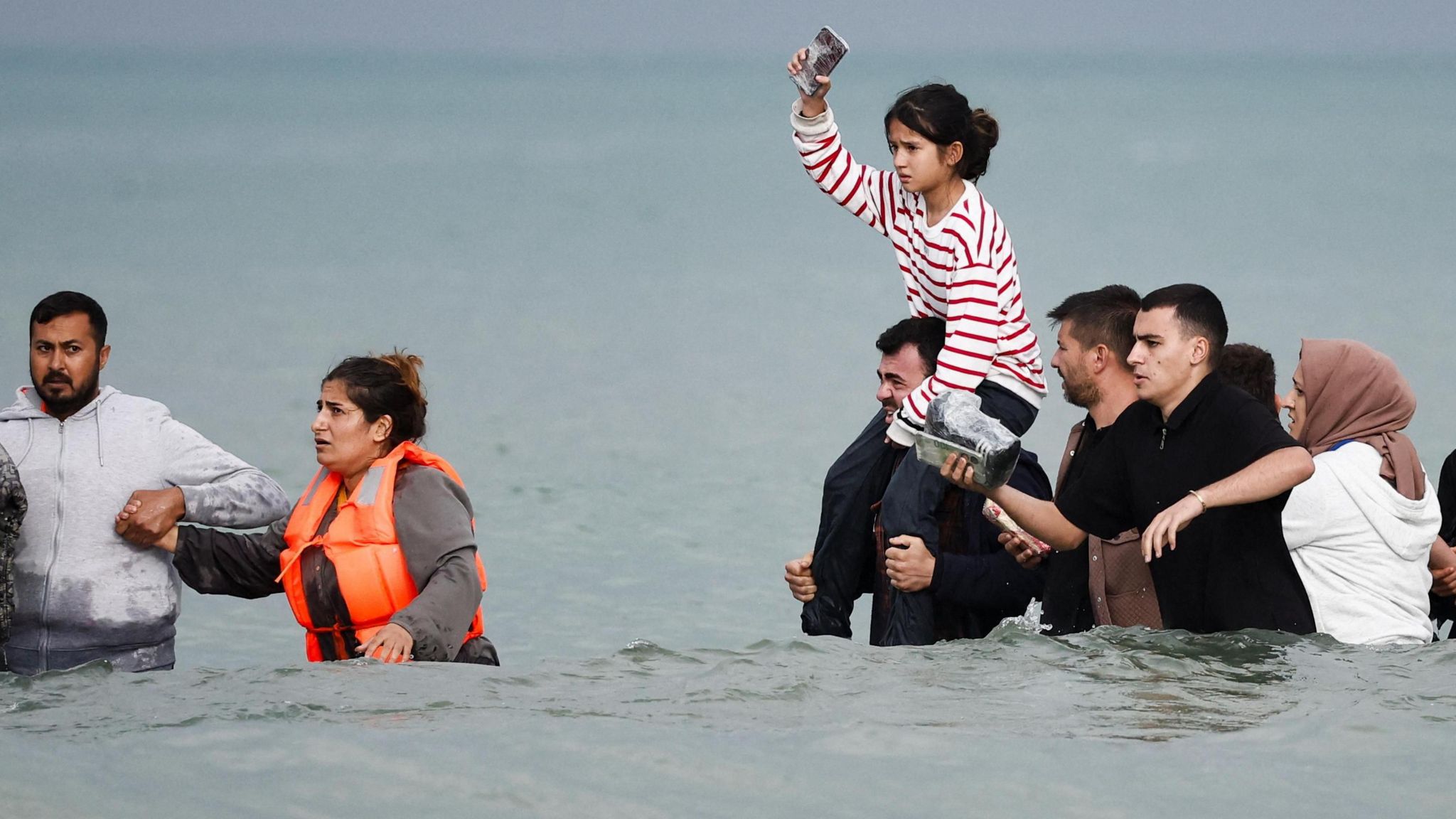 A group of people including women and a young girl on the shoulders of a man wade through waist-high sea water on their way to an inflatable boat.