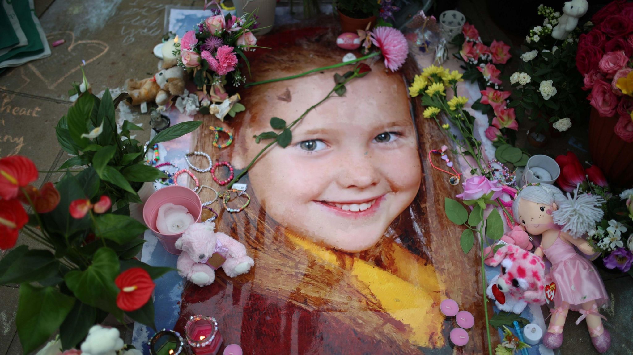 A large photograph of Elsie Dot Stancombe on the ground surrounded by flowers, candles, friendship bracelets and dolls.