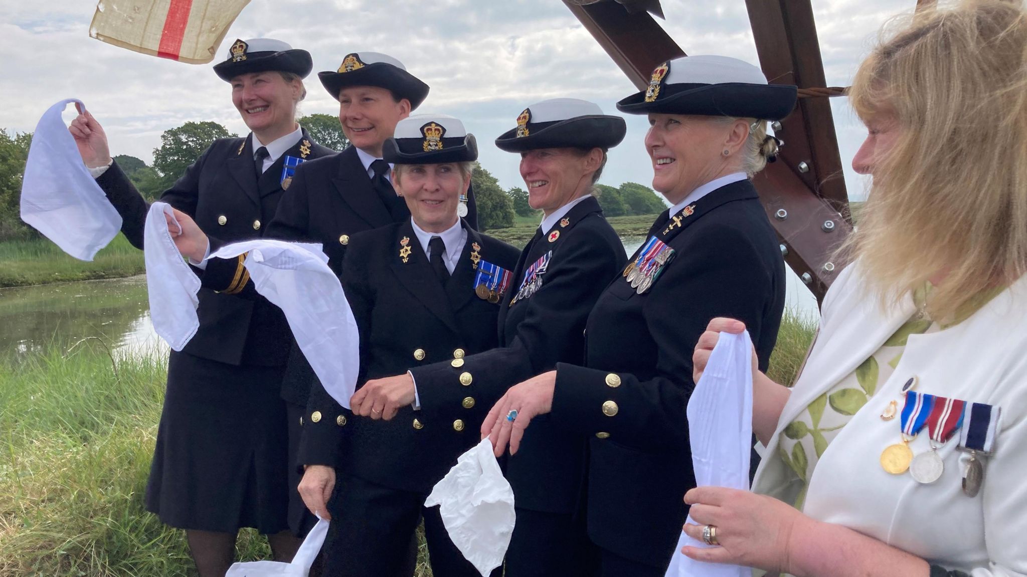 A group of six white women - five of which are wearing Royal Navy uniform, smile looking away from the camera and hold white handkerchiefs